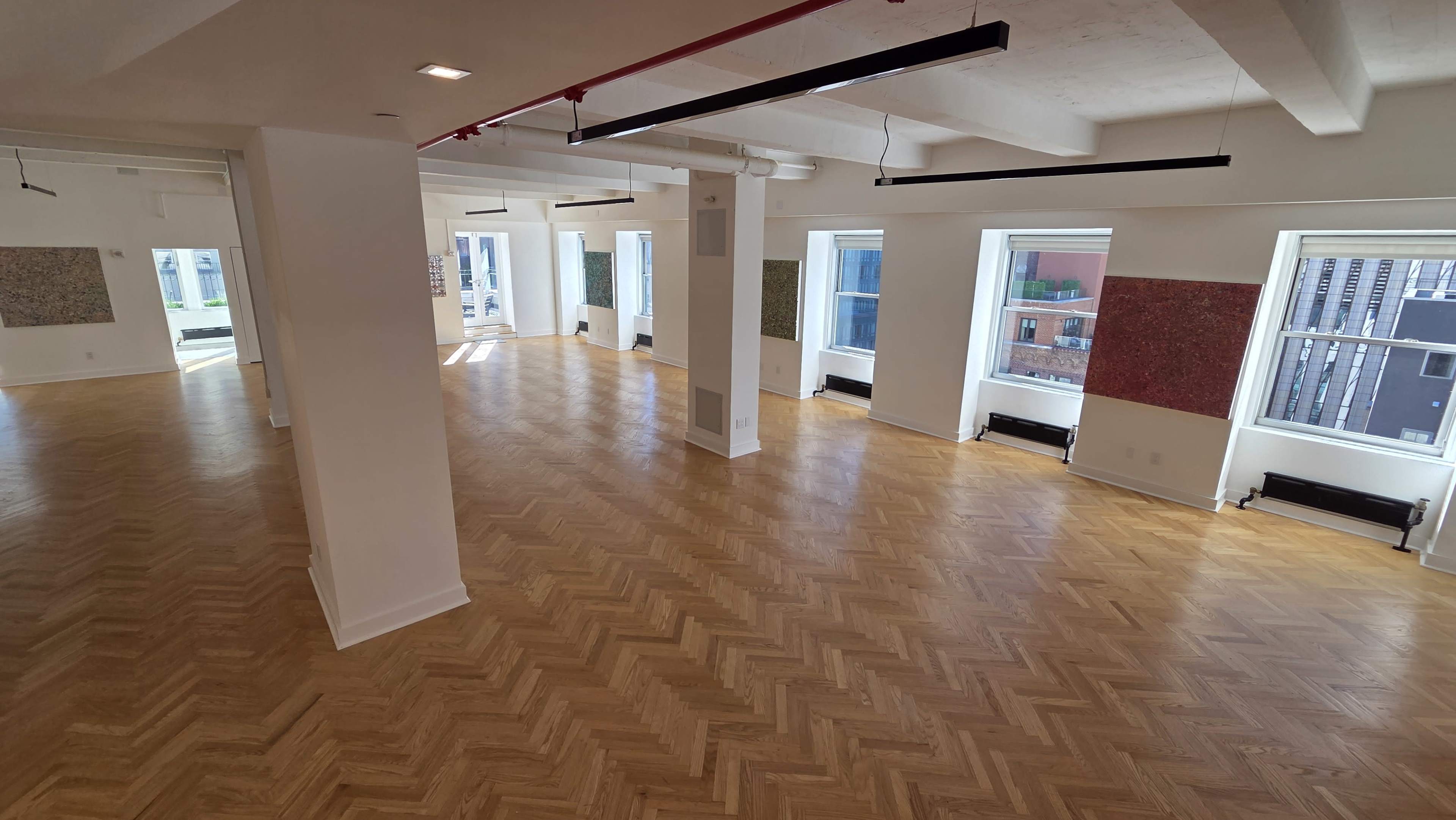 The image shows a large, empty room with wooden herringbone flooring, bright natural light coming through multiple windows, and art on the walls.