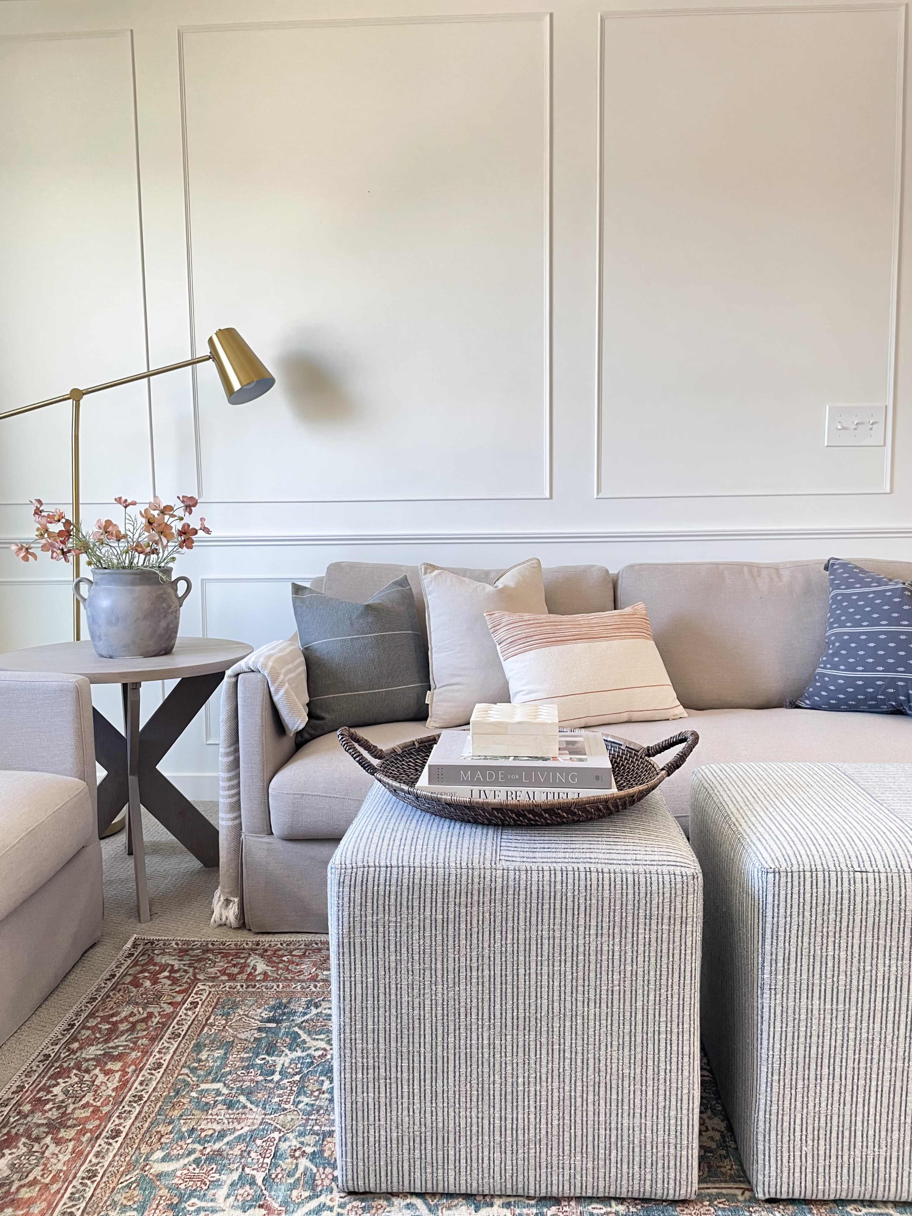 A light-colored living room features a gray sofa with various throw pillows, a woven tray holding books on a striped ottoman, and a floor lamp beside a vase of flowers.