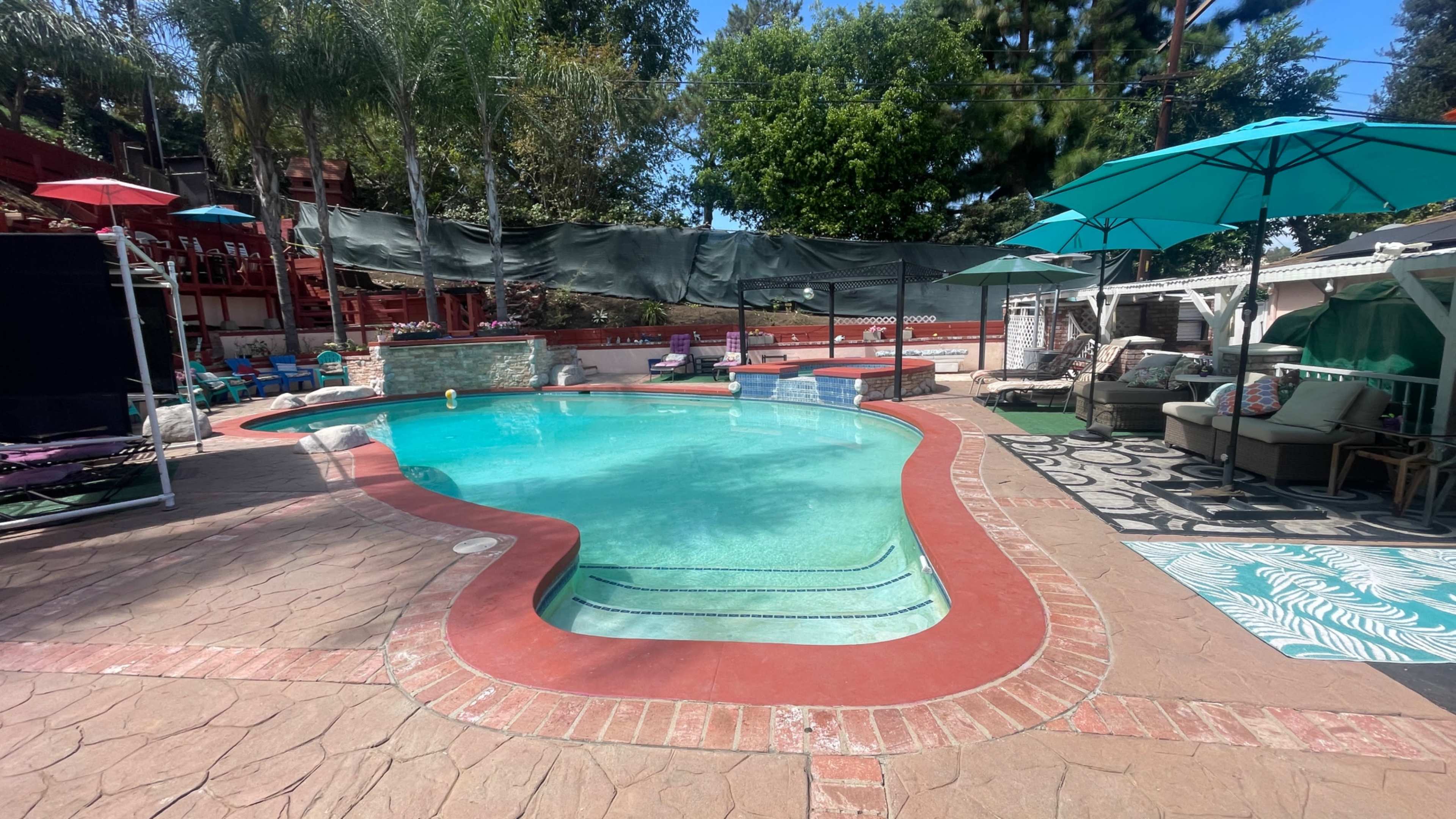 The image shows a swimming pool with a unique shape, surrounded by lounge chairs, umbrellas, and various outdoor furniture in a landscaped area.