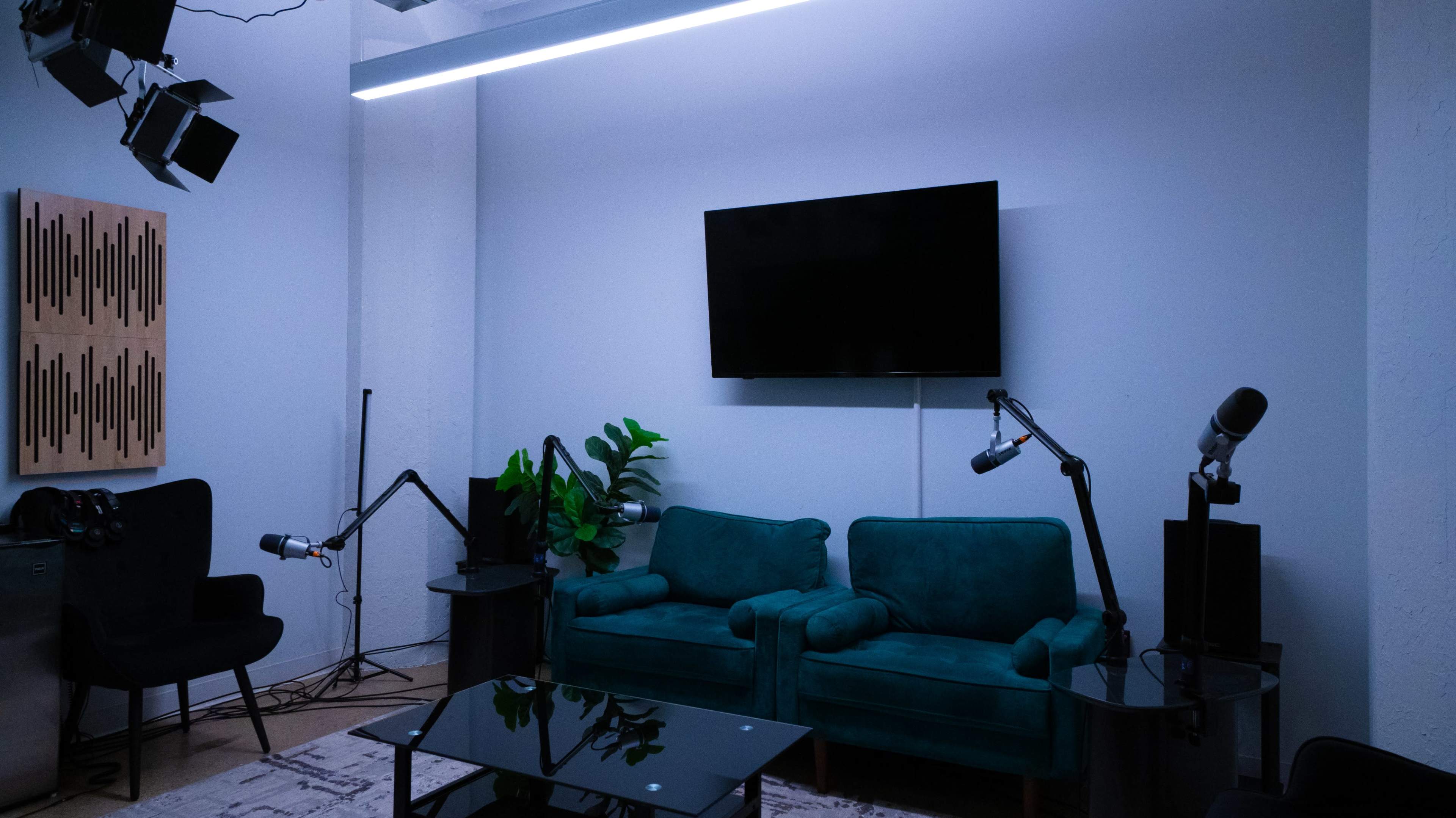 The image shows a modern studio setup with a large flat-screen TV on the wall, green sofas, a glass coffee table, and several microphones on stands.