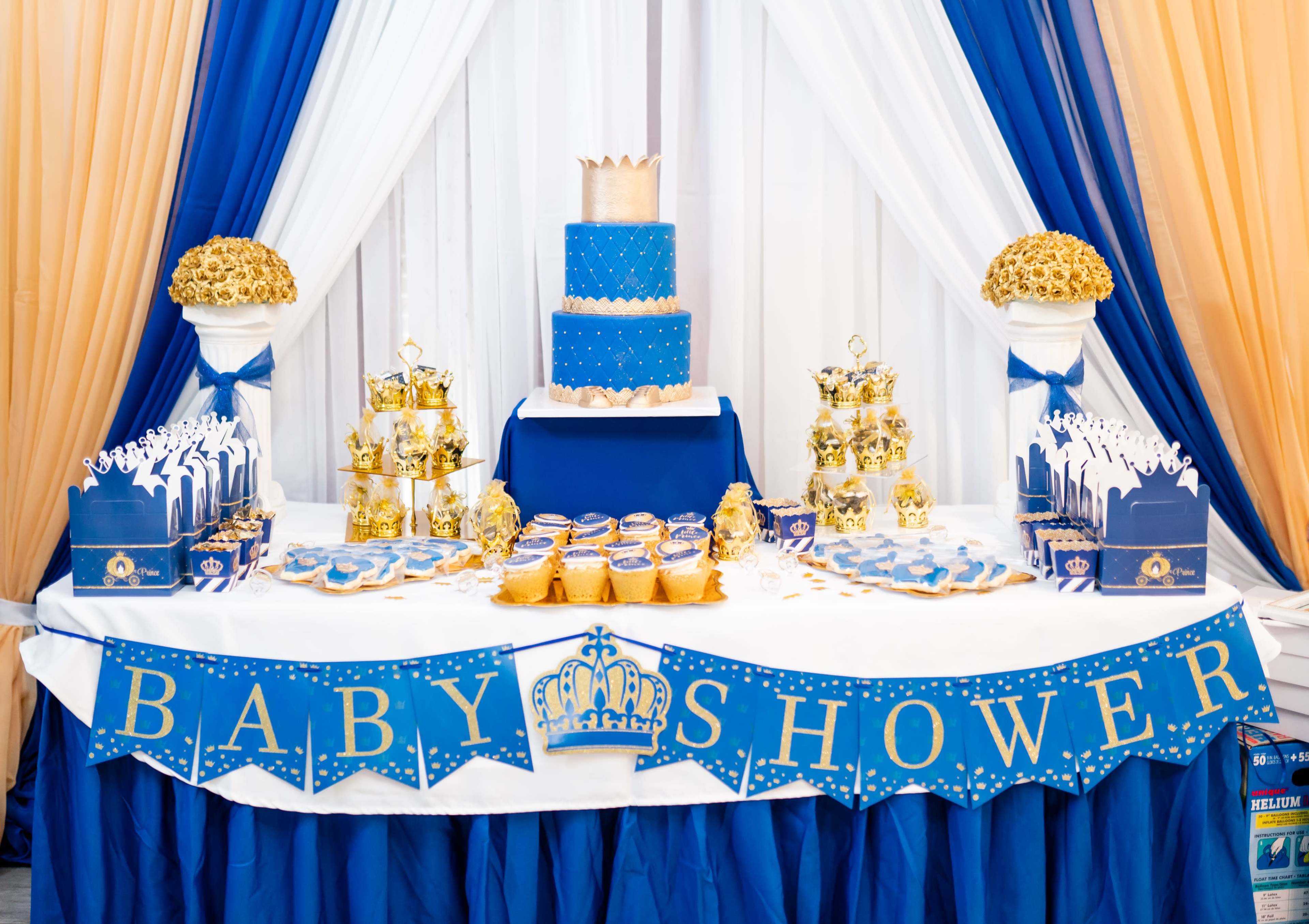 A decorated table features a tiered cake, golden treats, and a "BABY SHOWER" banner against a backdrop of blue and gold drapery.