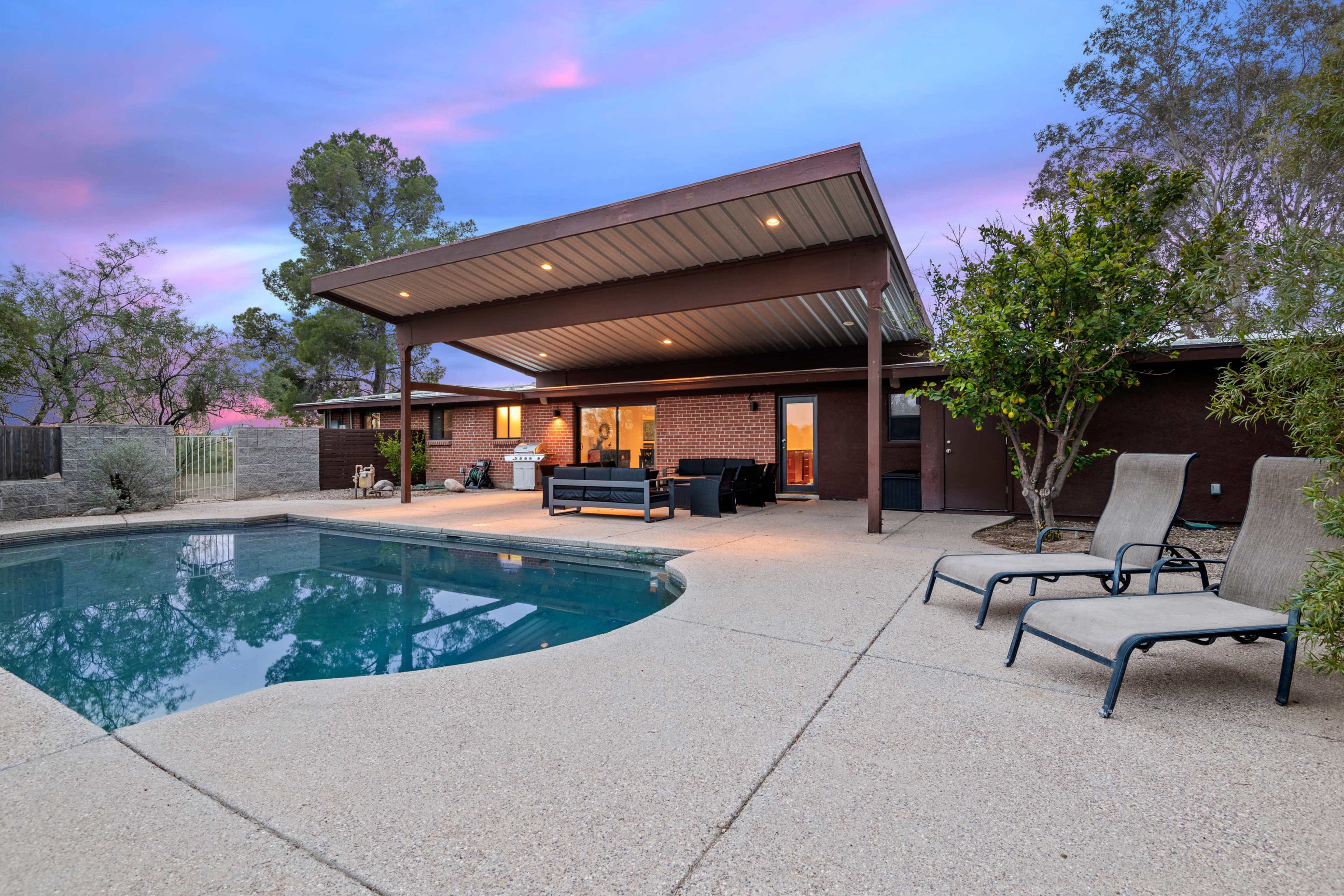 The image shows a backyard with a pool surrounded by patio furniture and a house with a covered area, set against a colorful sky at dusk.