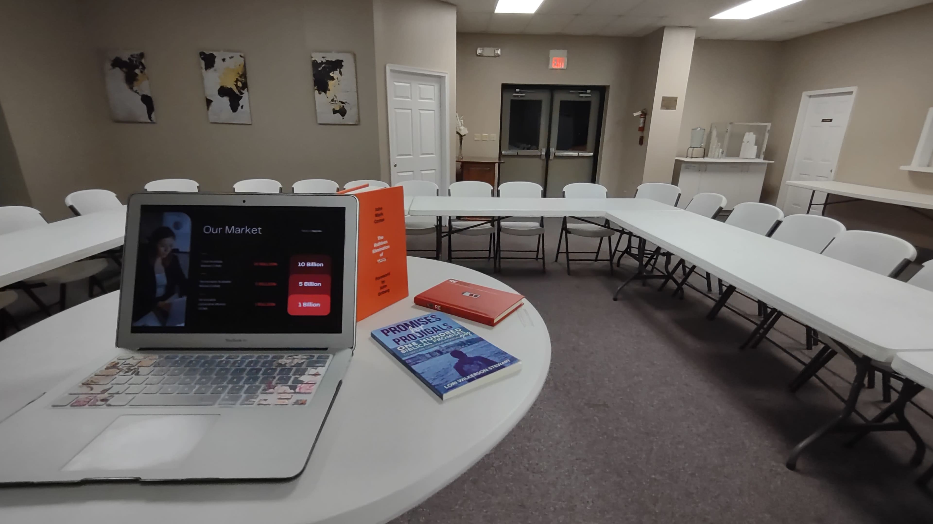 A laptop displaying a presentation is positioned on a table in a conference room with several rows of white chairs arranged around it.