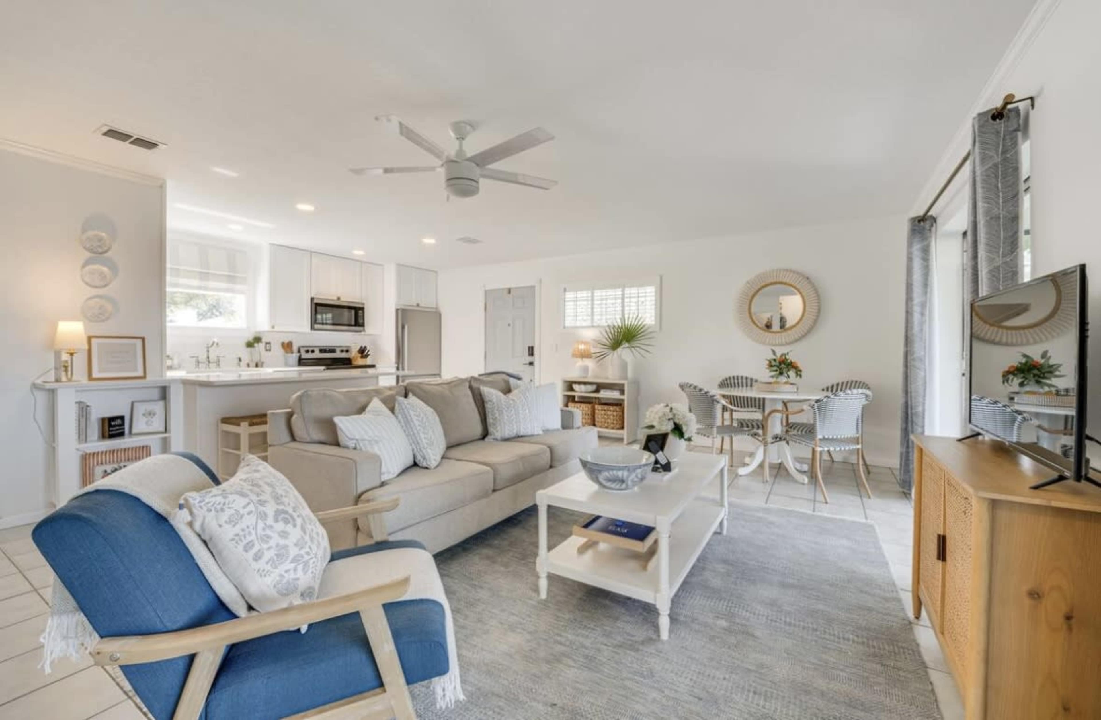 The image shows a bright living room with a beige sofa, a blue accent chair, and a white coffee table, adjacent to an open kitchen.