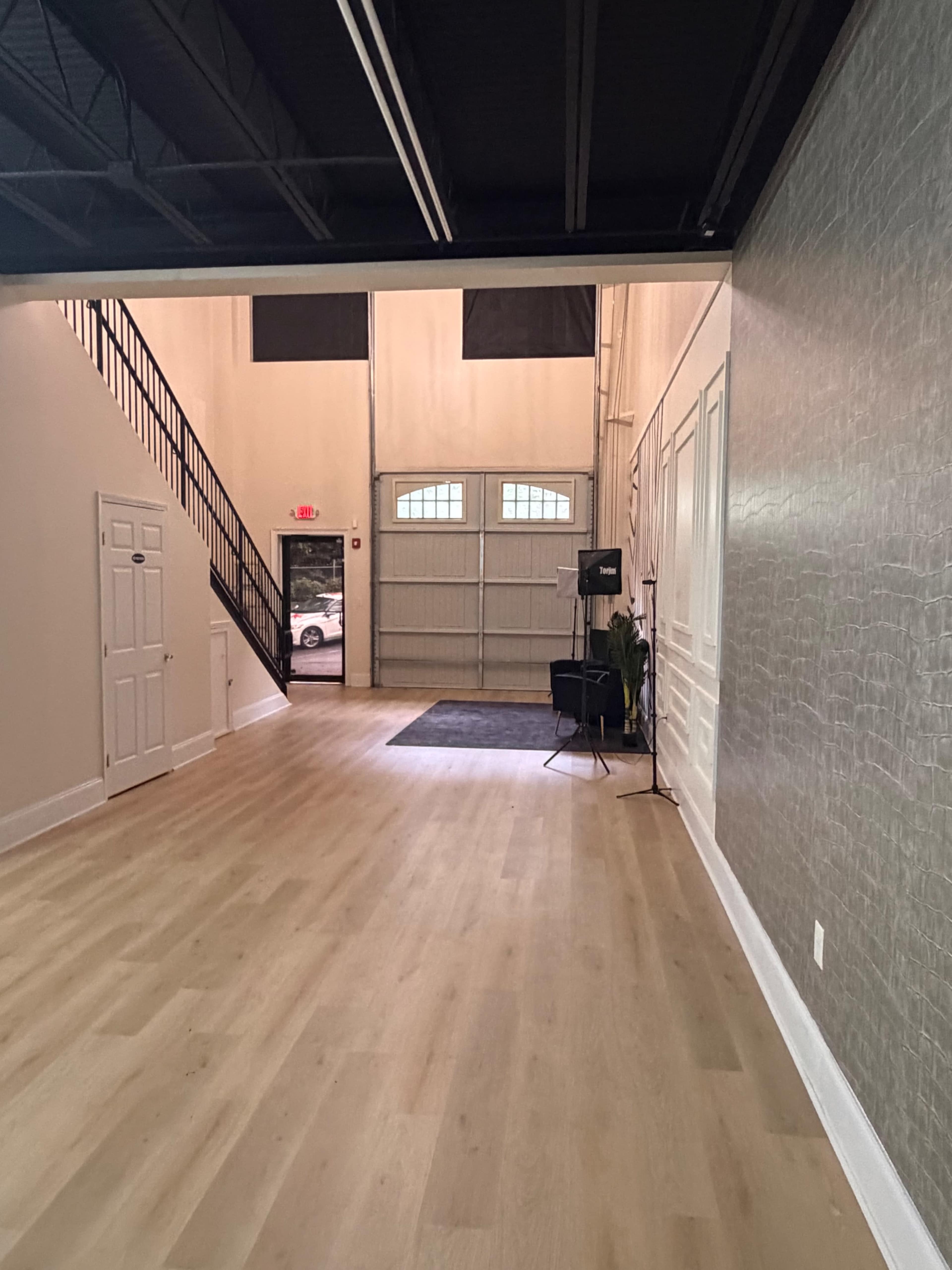 The image shows an empty open space with wooden flooring, featuring a staircase on the left, a large garage door at the back, and a small seating area to the right.