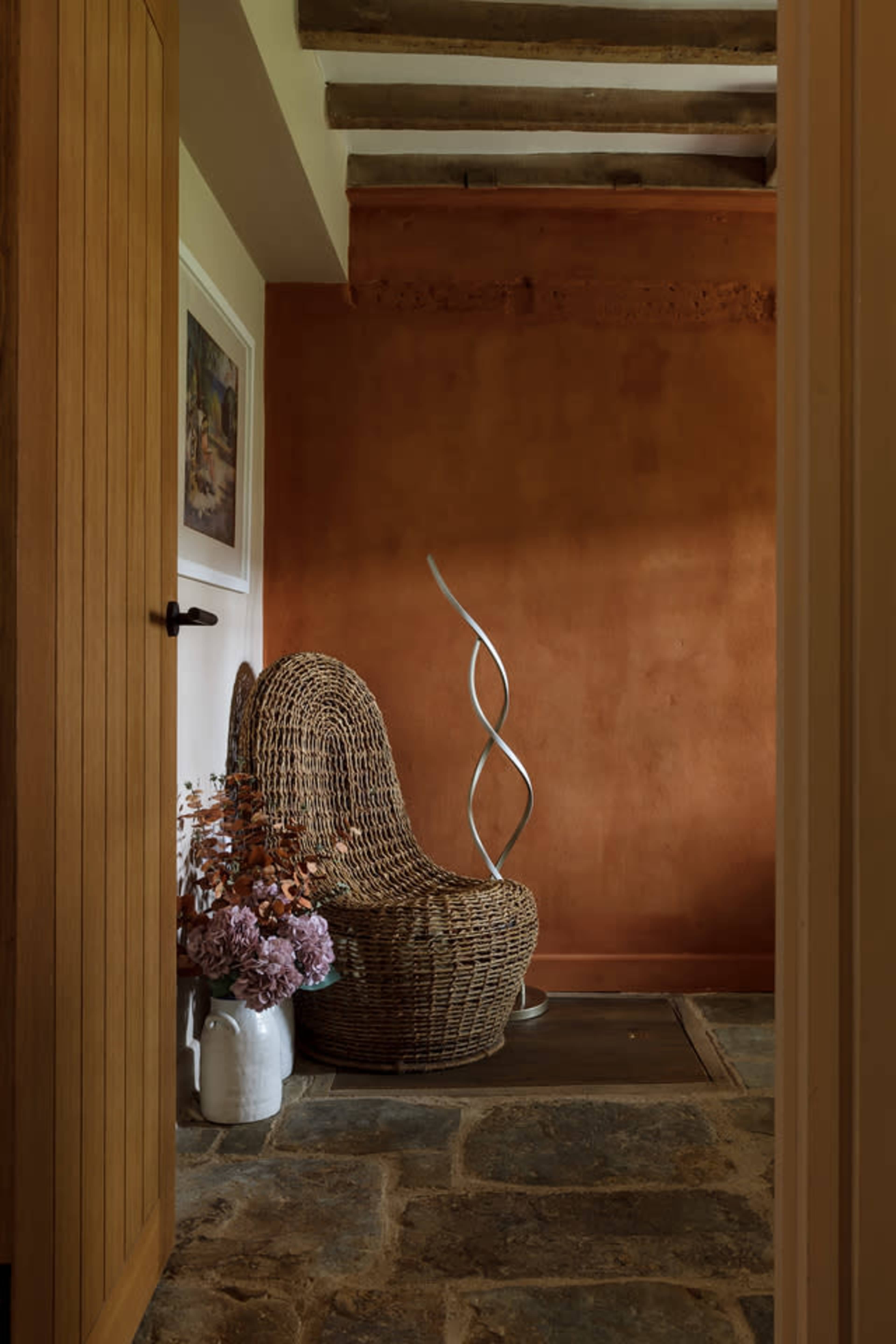 A wicker chair is positioned against an orange wall, with a modern sculpture and a vase of dried flowers nearby, all seen through an open doorway.