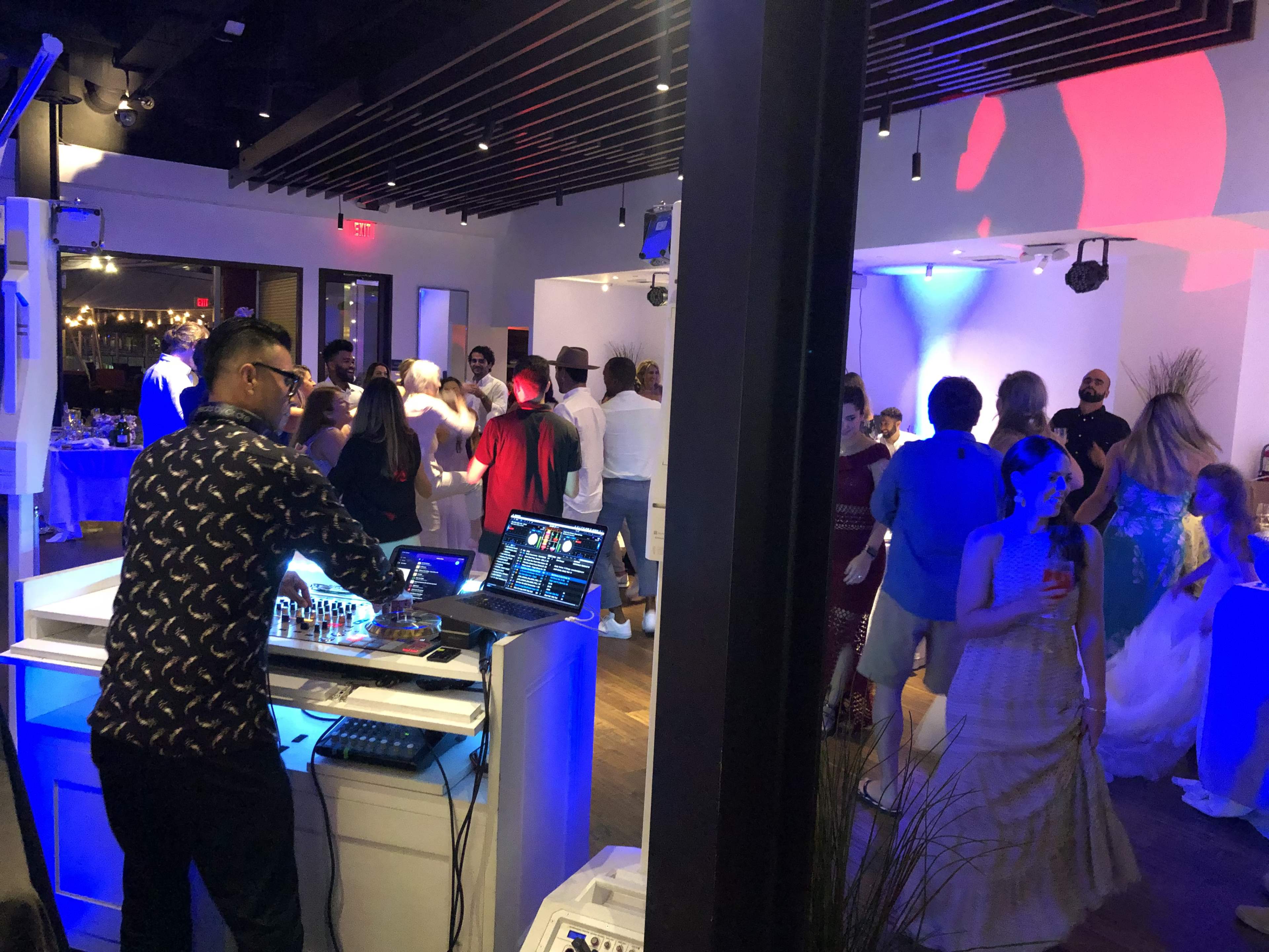 A DJ mixes music at a lively dance party in a modern venue filled with guests dancing and socializing.