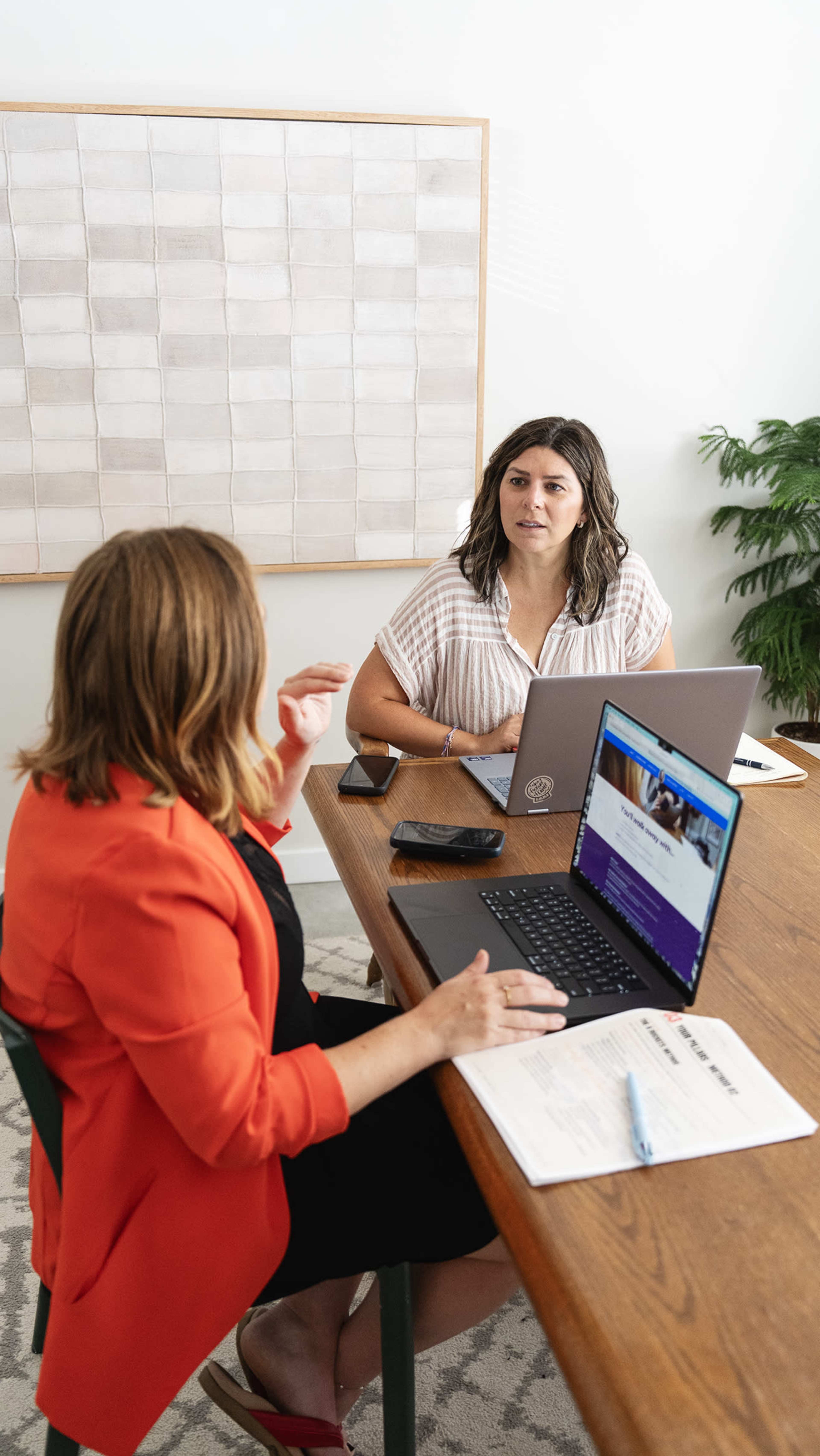 Two women are seated at a wooden table, engaged in discussion while using laptops and a smartphone in a well-lit office setting.