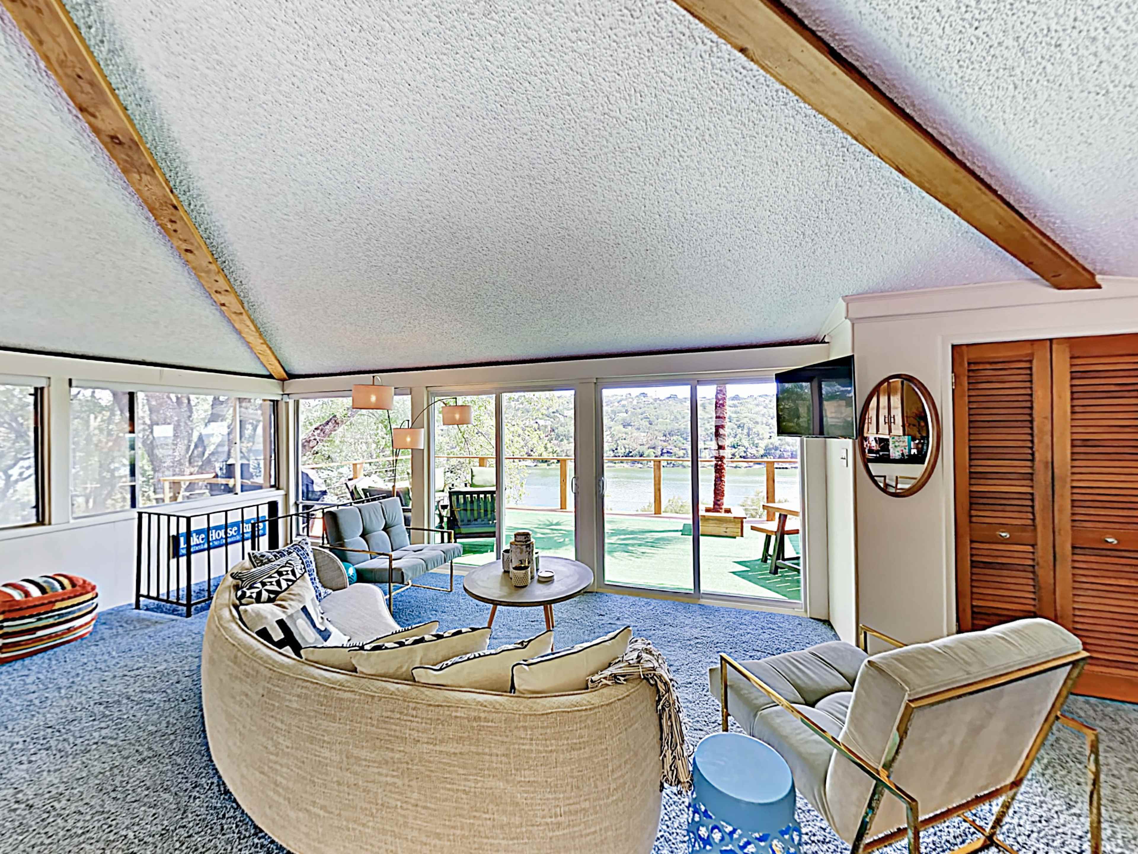 The image shows a living room with large windows overlooking a balcony, featuring a curved sofa, matching armchairs, and a small coffee table.