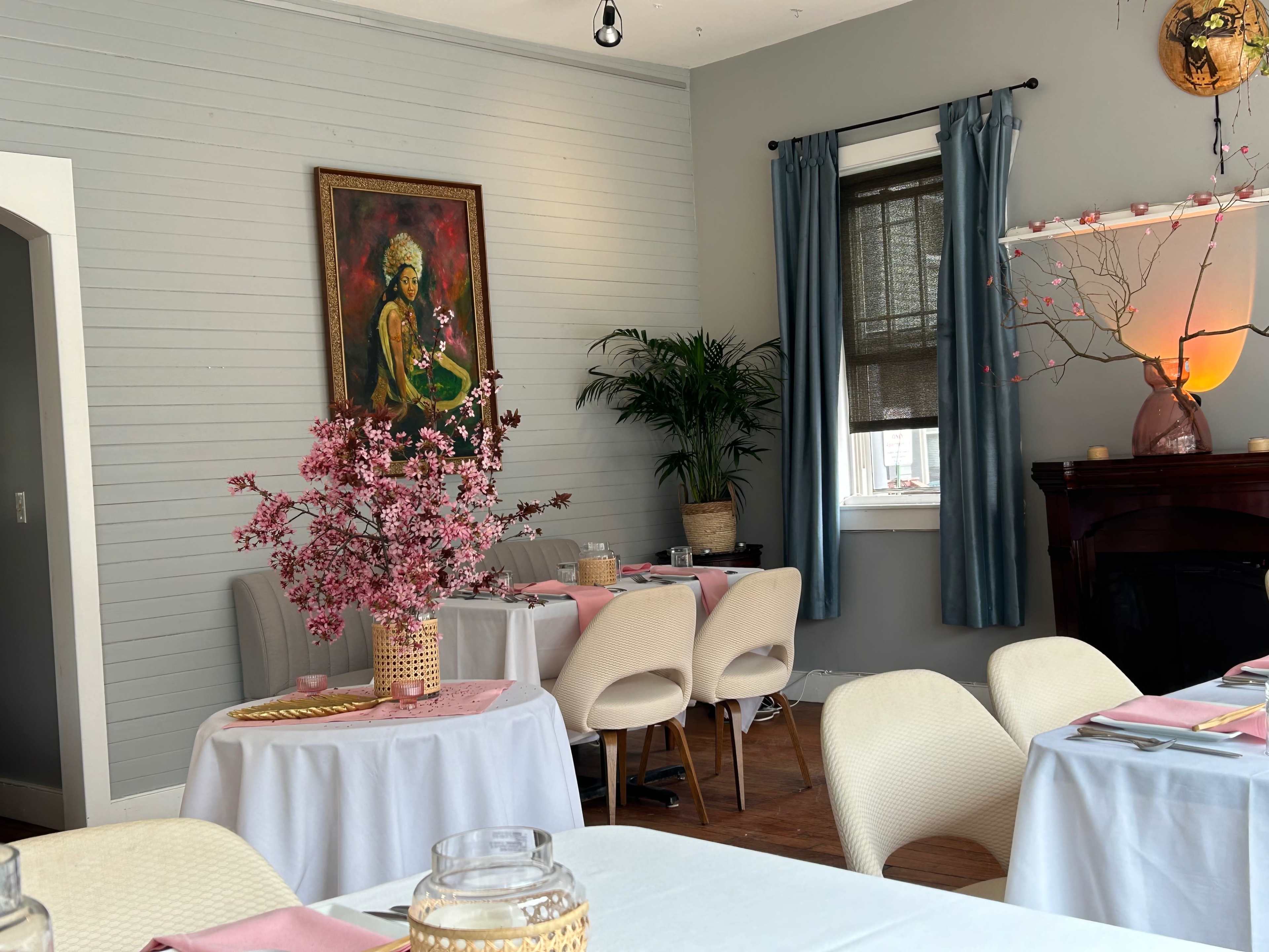 A dining area with white tables, a potted plant, and a large piece of artwork on the wall, complemented by floral centerpieces and light blue curtains.