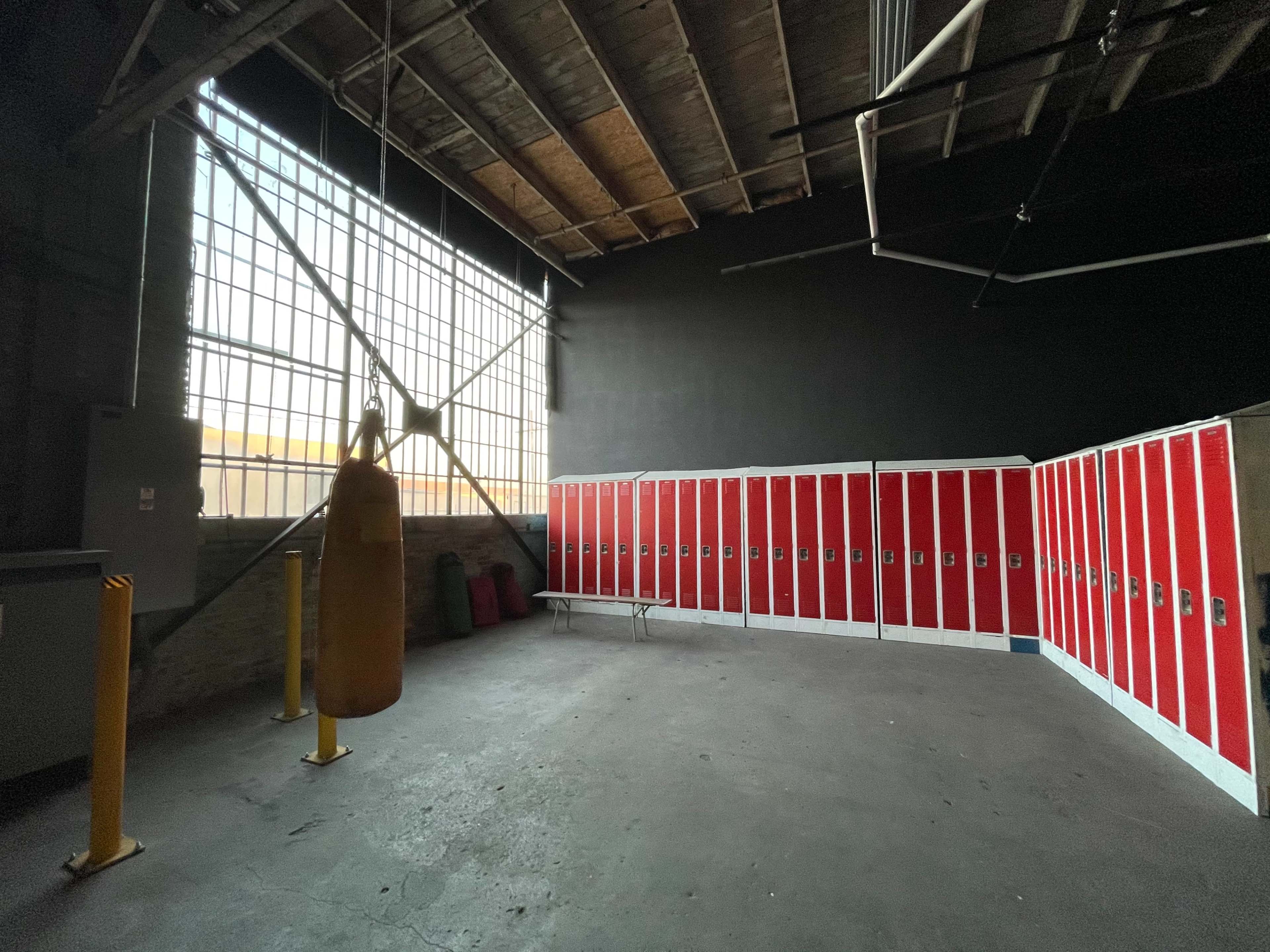 The image shows a sparsely decorated gym interior featuring a heavy punching bag suspended from the ceiling and a row of red lockers along one wall.
