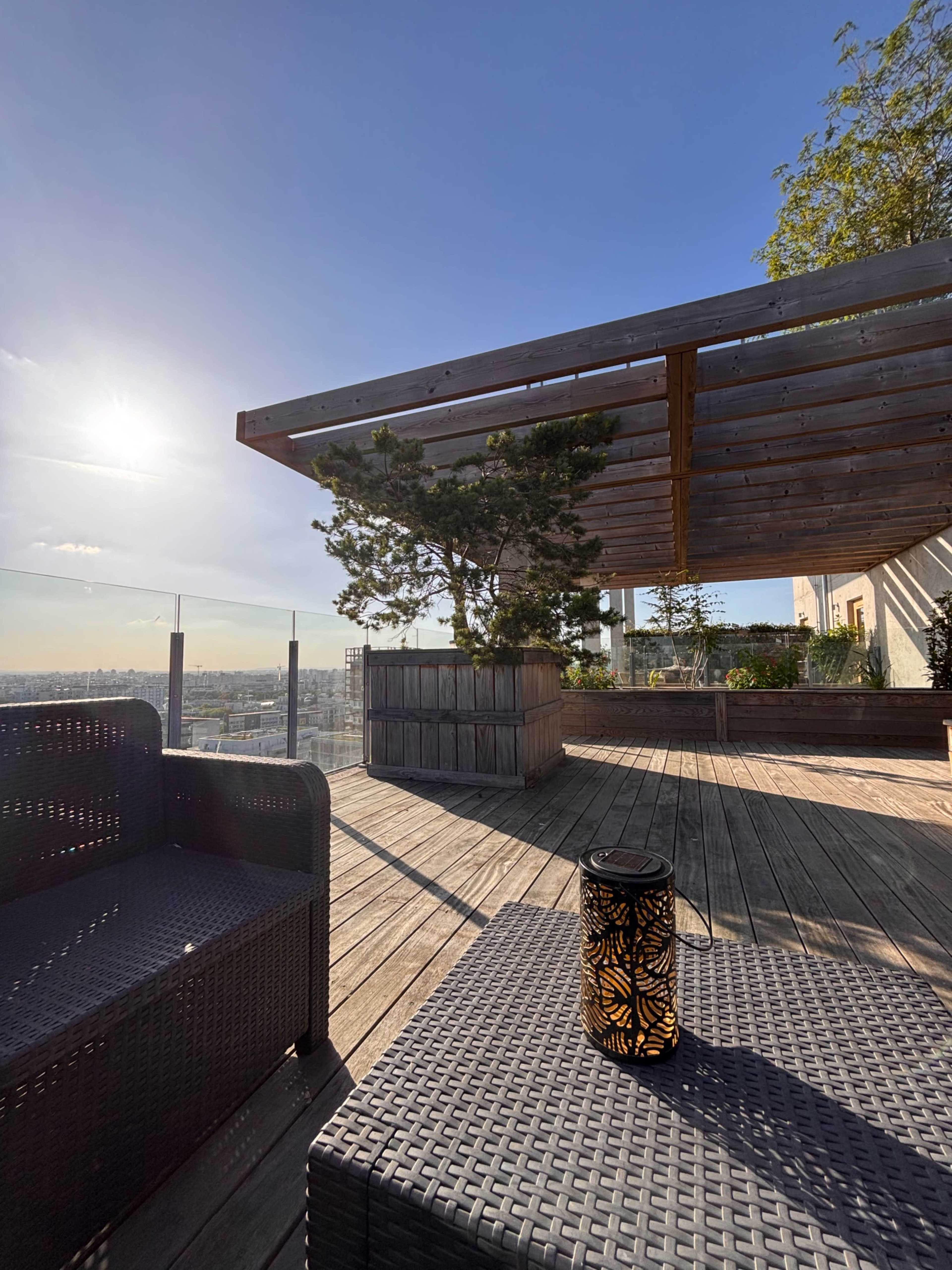 A rooftop terrace features outdoor furniture, wooden decking, and a lantern, with a view of the skyline under clear blue skies.