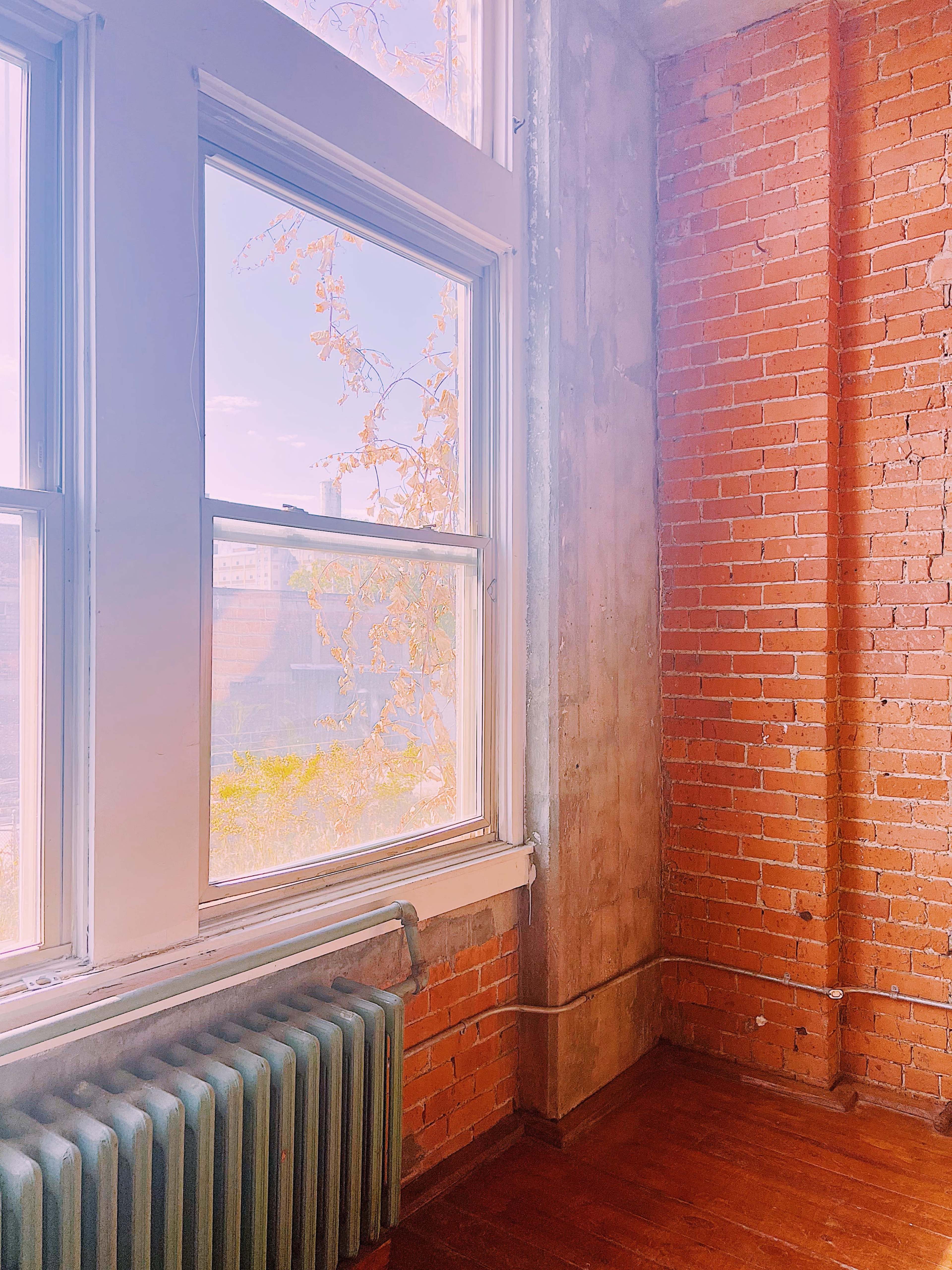 A corner of a room with exposed brick walls, a large window, and a vintage radiator beneath the window.