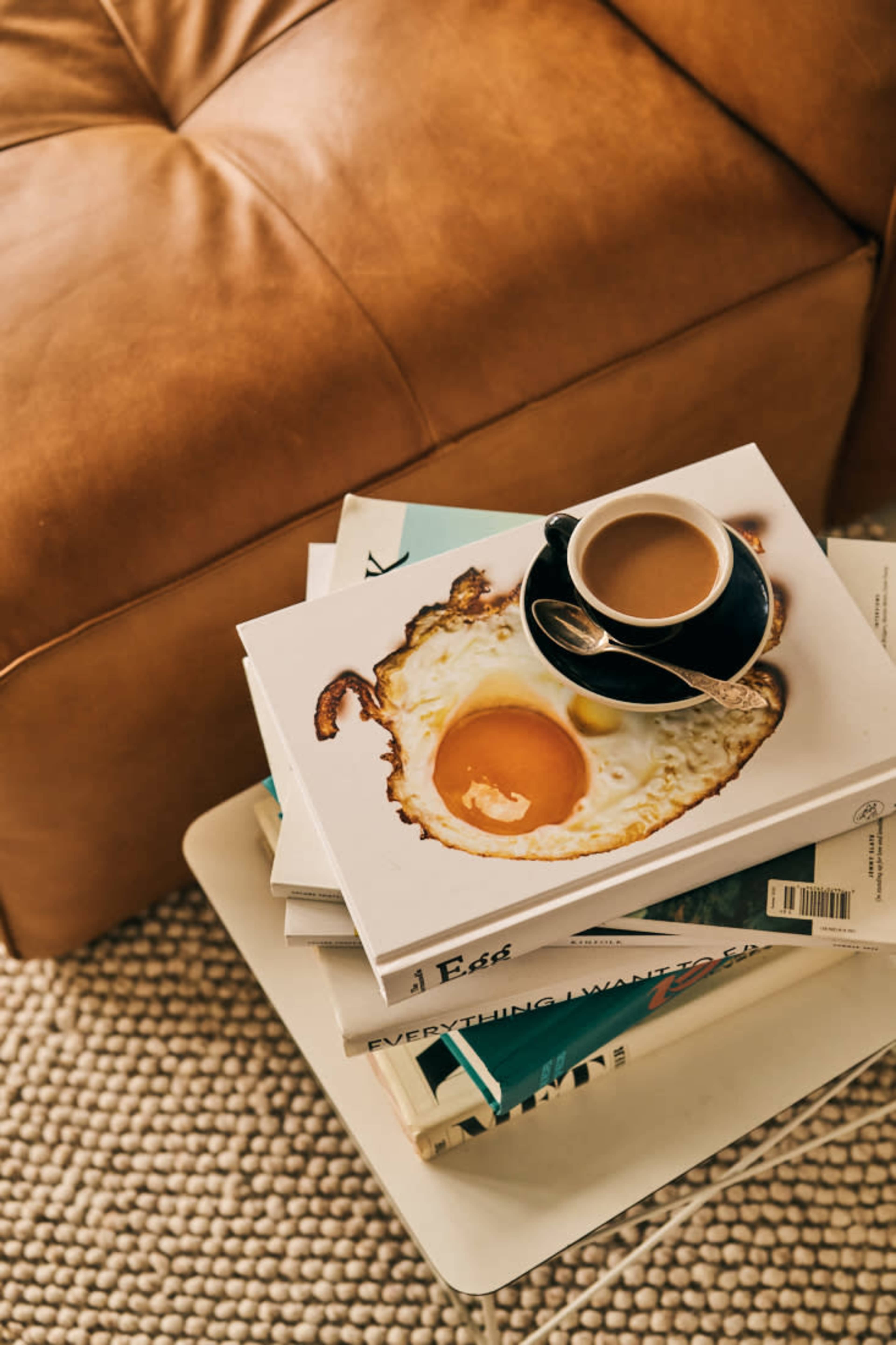 A black cup of coffee sits on a stack of books topped with a cookbook featuring a fried egg on its cover, next to a leather sofa.