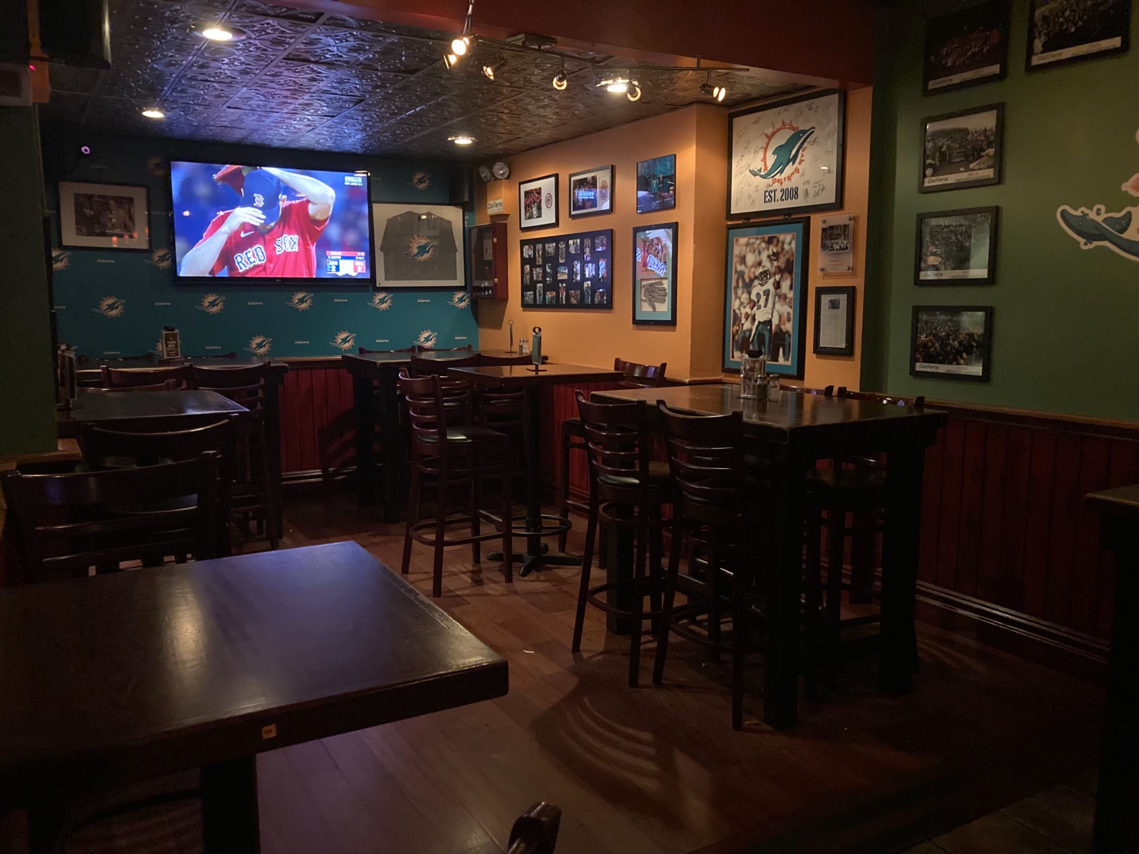 The image shows the interior of a bar with wooden tables and chairs, featuring a large screen displaying a sports game and walls adorned with framed photographs.