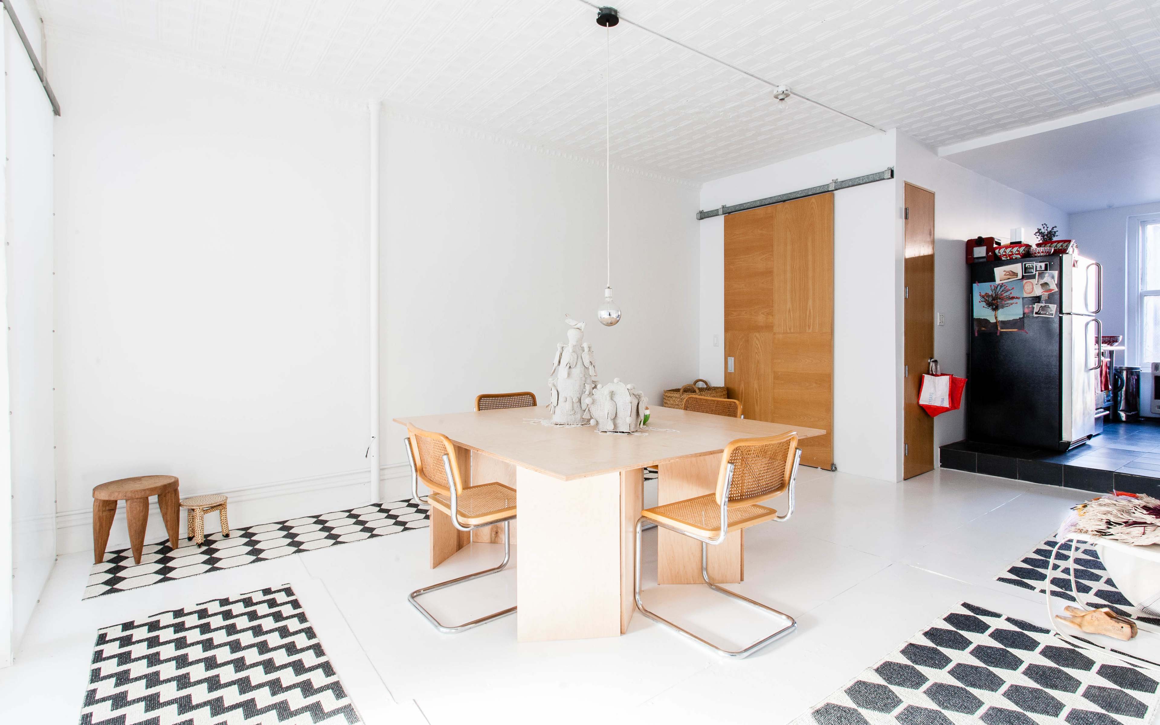 A minimalist dining area with a square wooden table surrounded by four woven chairs, accented by patterned black and white rugs.