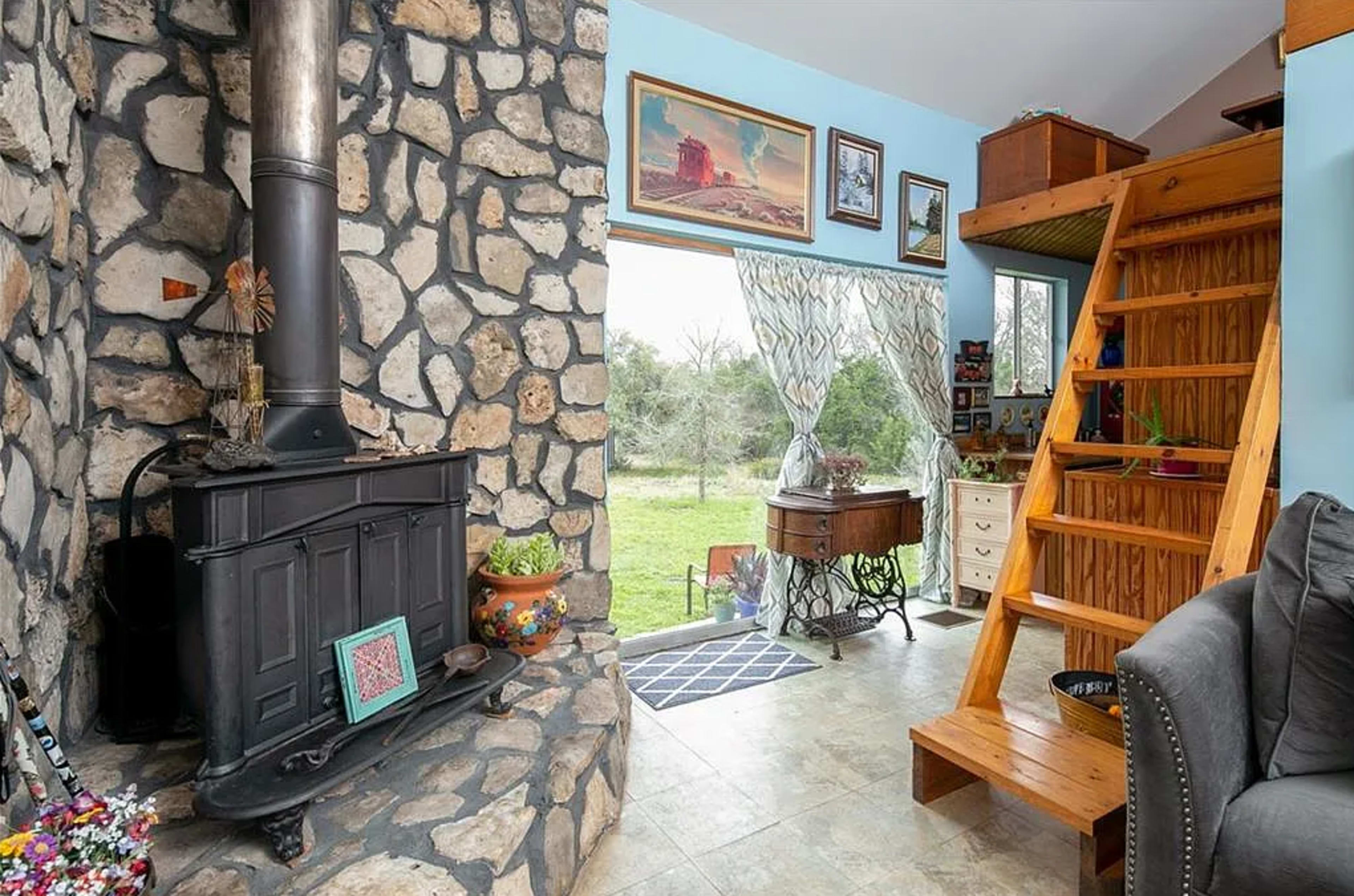 The image shows a living space featuring a stone fireplace, a wooden staircase leading to a loft, and large windows with sheer curtains allowing light to enter.
