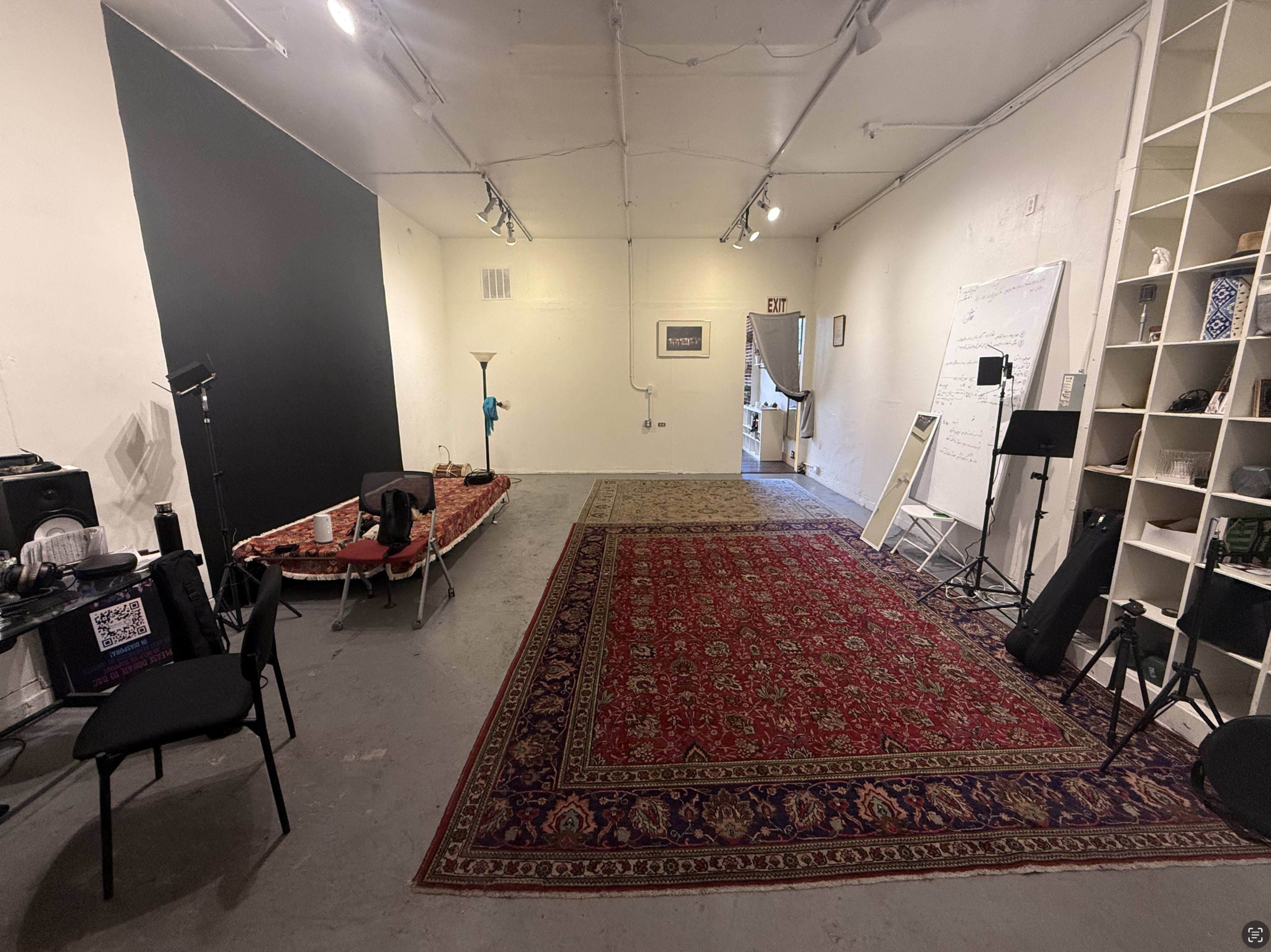 The image shows a spacious, well-lit room with a large area rug, a black wall section, and various furniture items including chairs and a lamp.