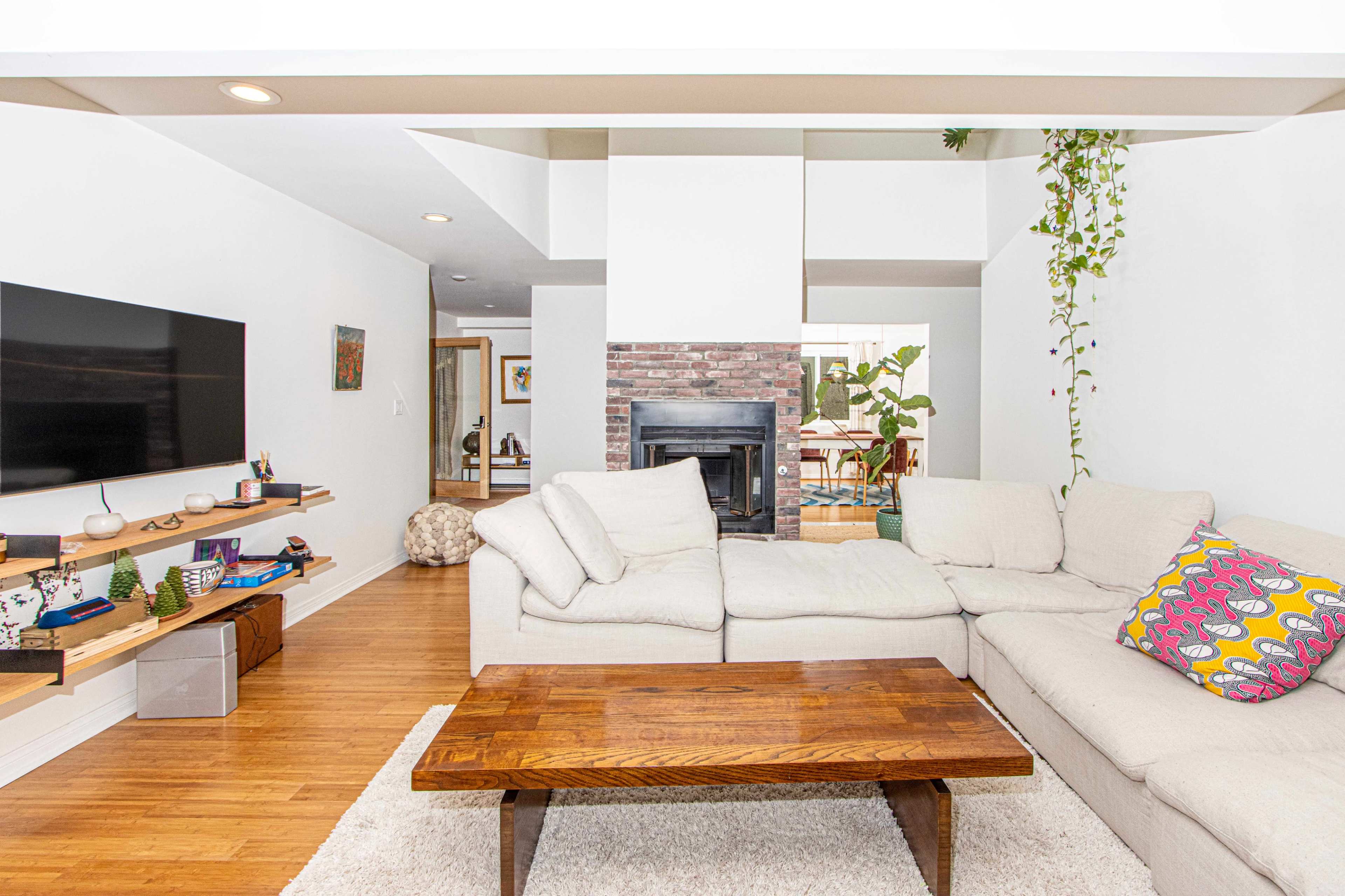 The image shows a modern living room featuring a light-colored sectional sofa, a wooden coffee table, and a brick fireplace, with a television mounted on the wall and greenery in the corner.