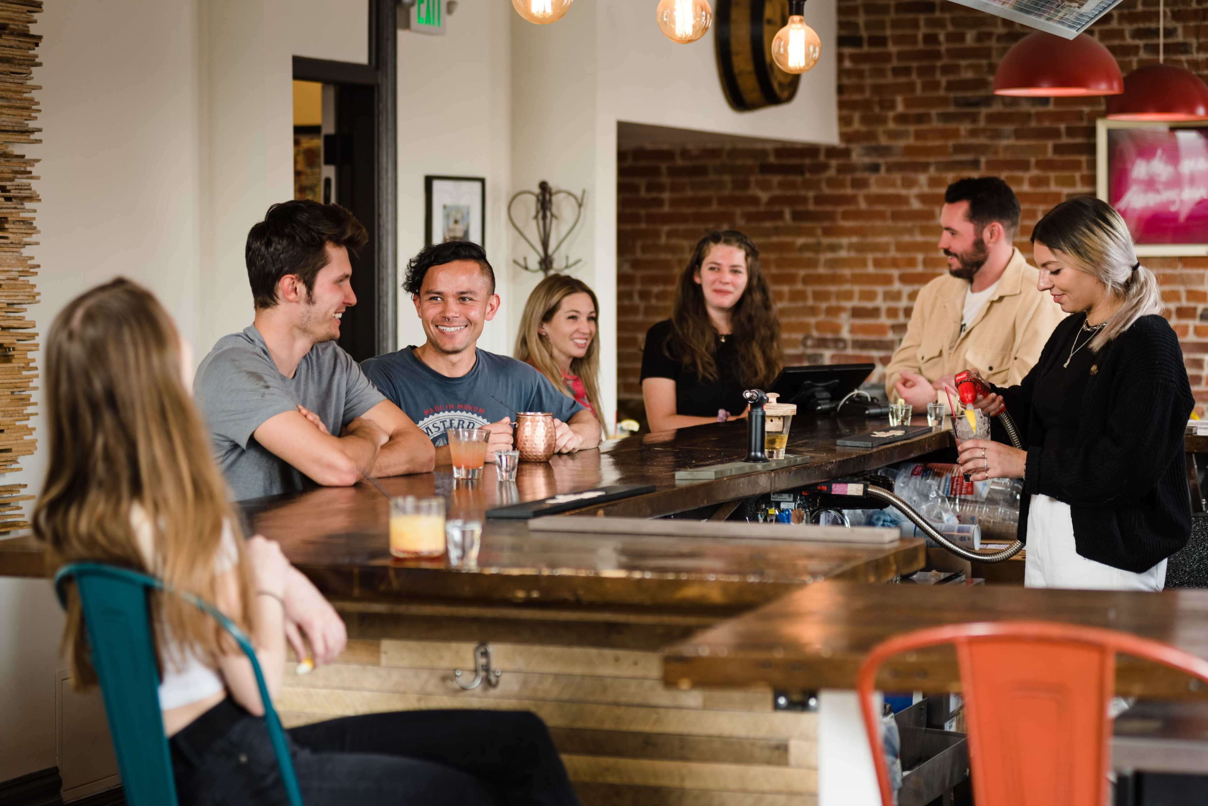 A group of people is gathered at a bar, interacting and enjoying drinks in a warmly lit space with exposed brick walls.