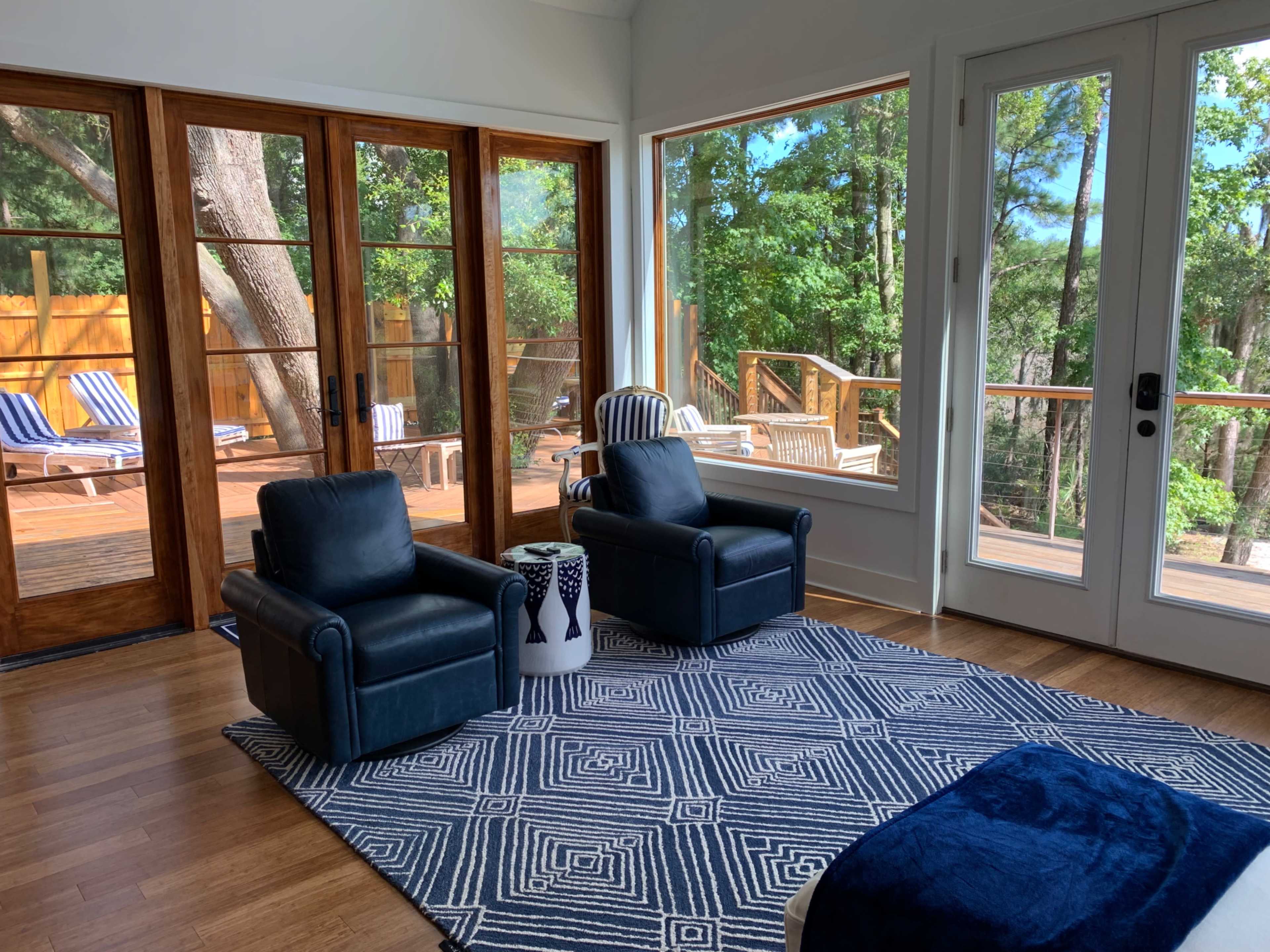 The image shows a modern living room with two black armchairs, a geometric patterned rug, and large windows overlooking a wooden deck and surrounding trees.
