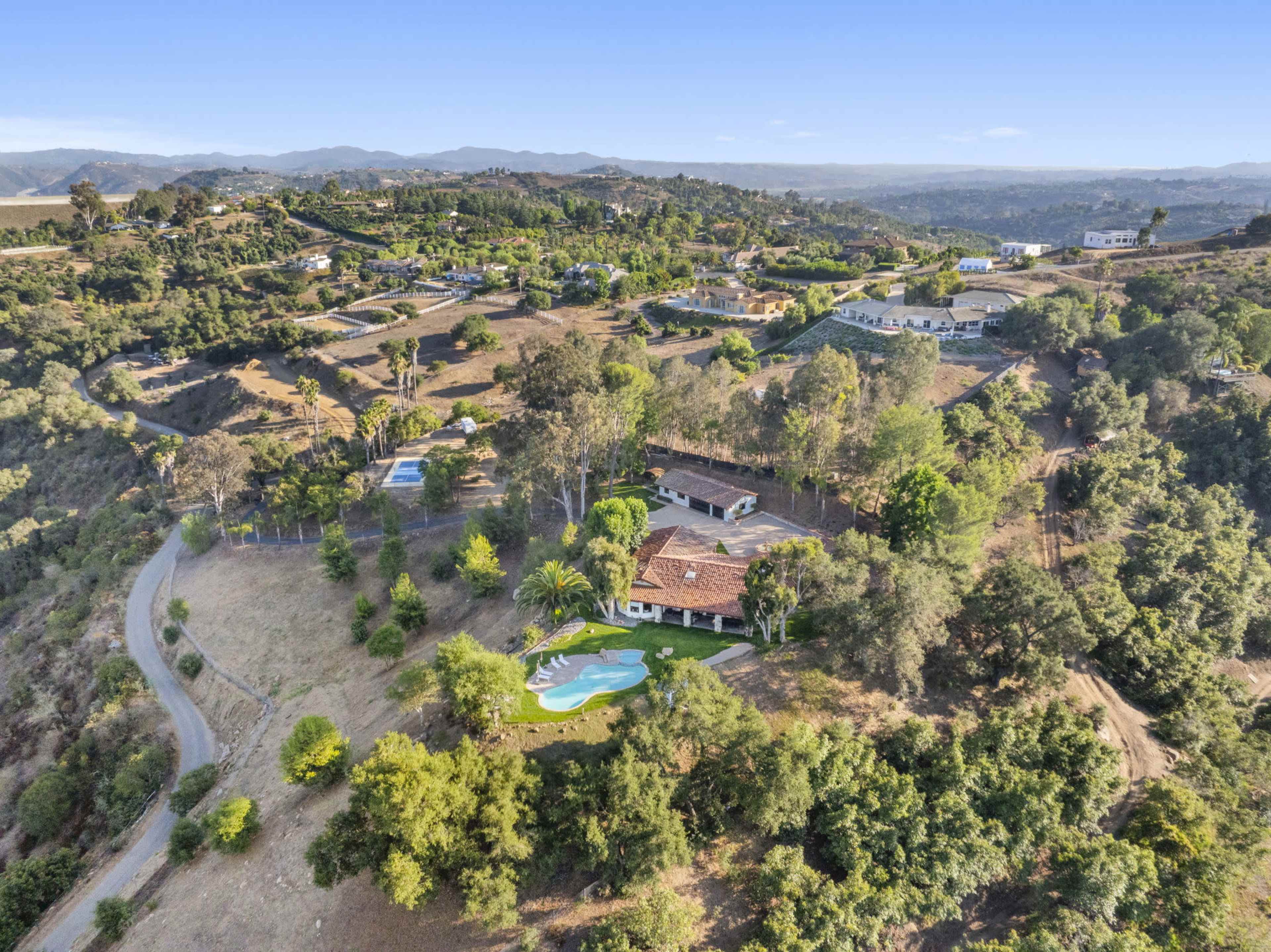 The image shows a sprawling estate with a pool surrounded by lush greenery, situated on a hillside with distant mountains in the background.