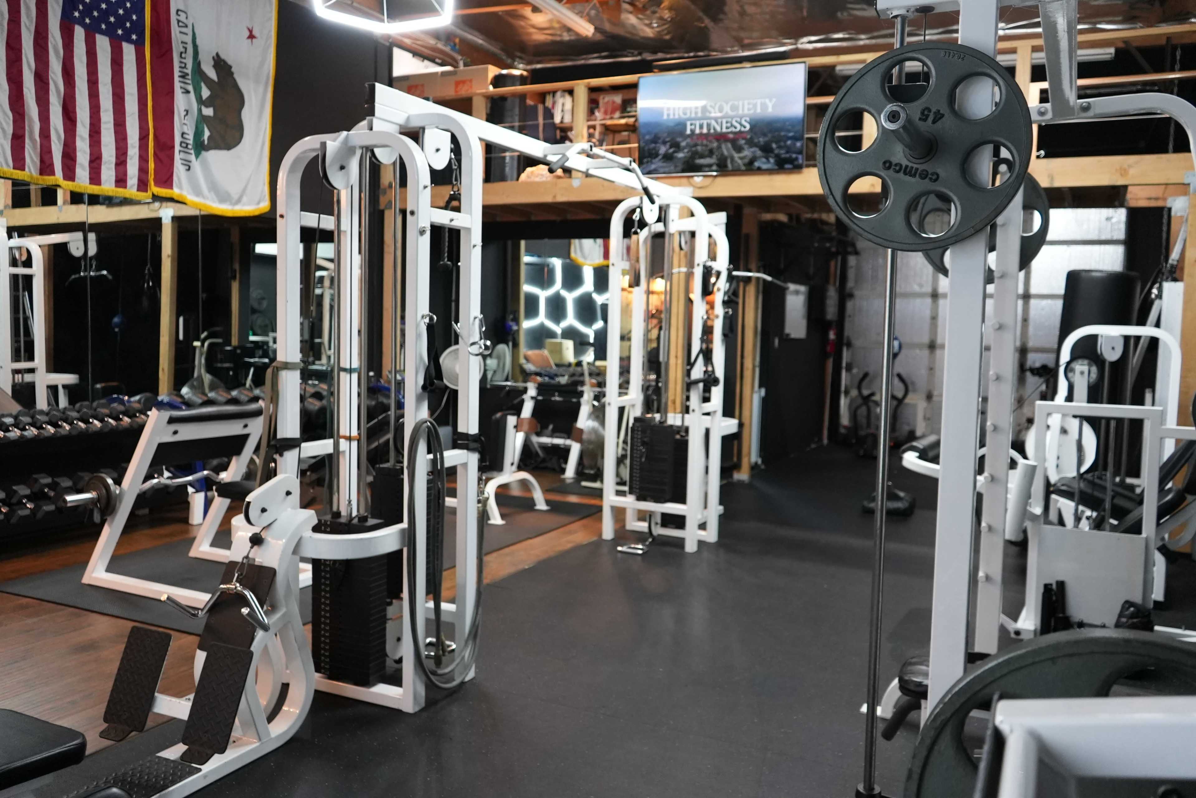 A well-equipped gym featuring various weight training machines, dumbbells, and a mirrored wall, with flags and a sign visible in the background.