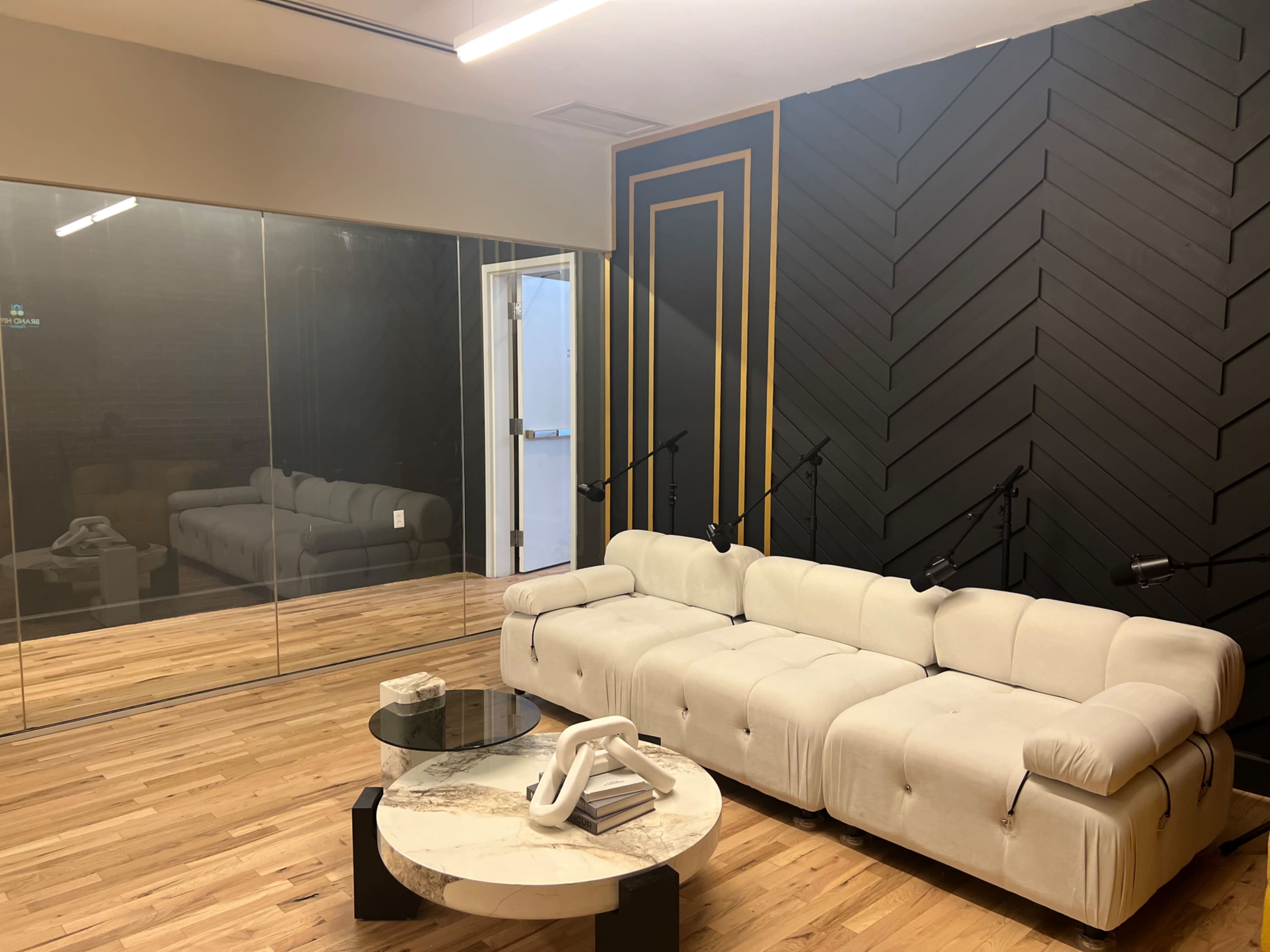 A modern lounge area featuring a beige sectional sofa, a marble coffee table, and a glass wall with a dark geometric-patterned accent wall behind it.