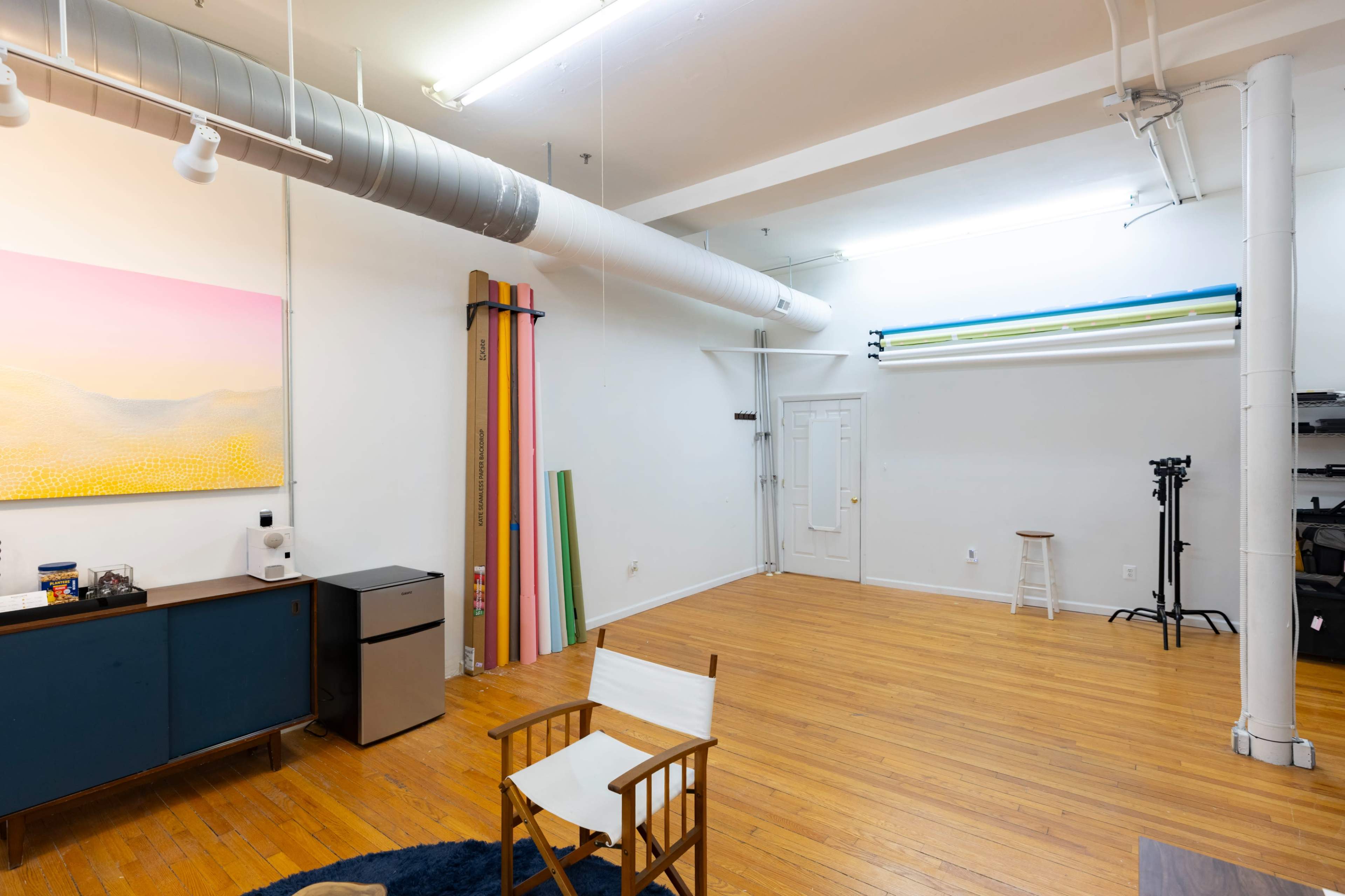 The image shows a spacious, well-lit studio featuring wooden floors, a white wall with colorful backdrops, and a few pieces of furniture including a chair and a small refrigerator.