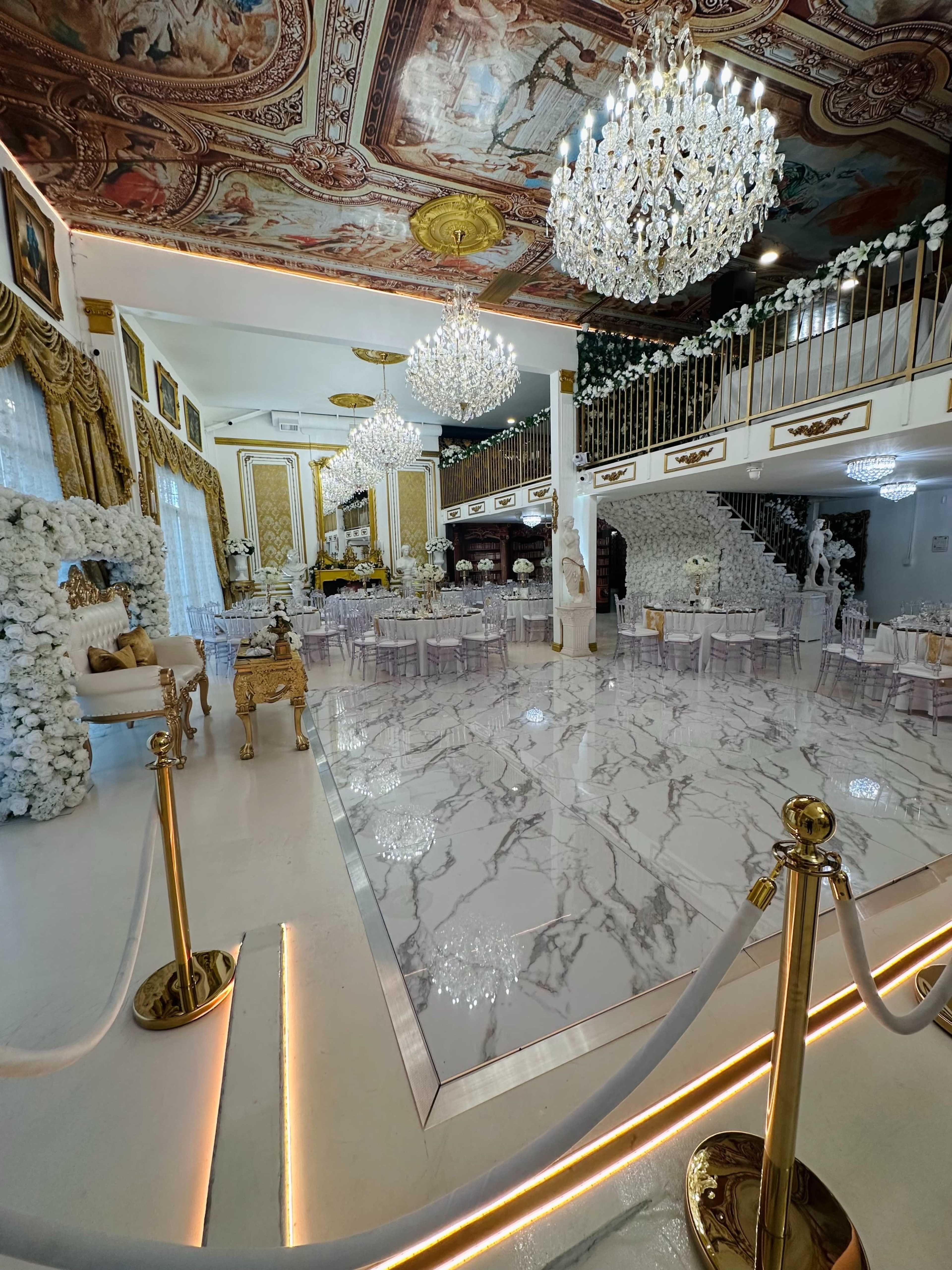 The image shows an opulent event space with marble flooring, crystal chandeliers, and elegantly decorated walls, featuring a staircase and floral arrangements.
