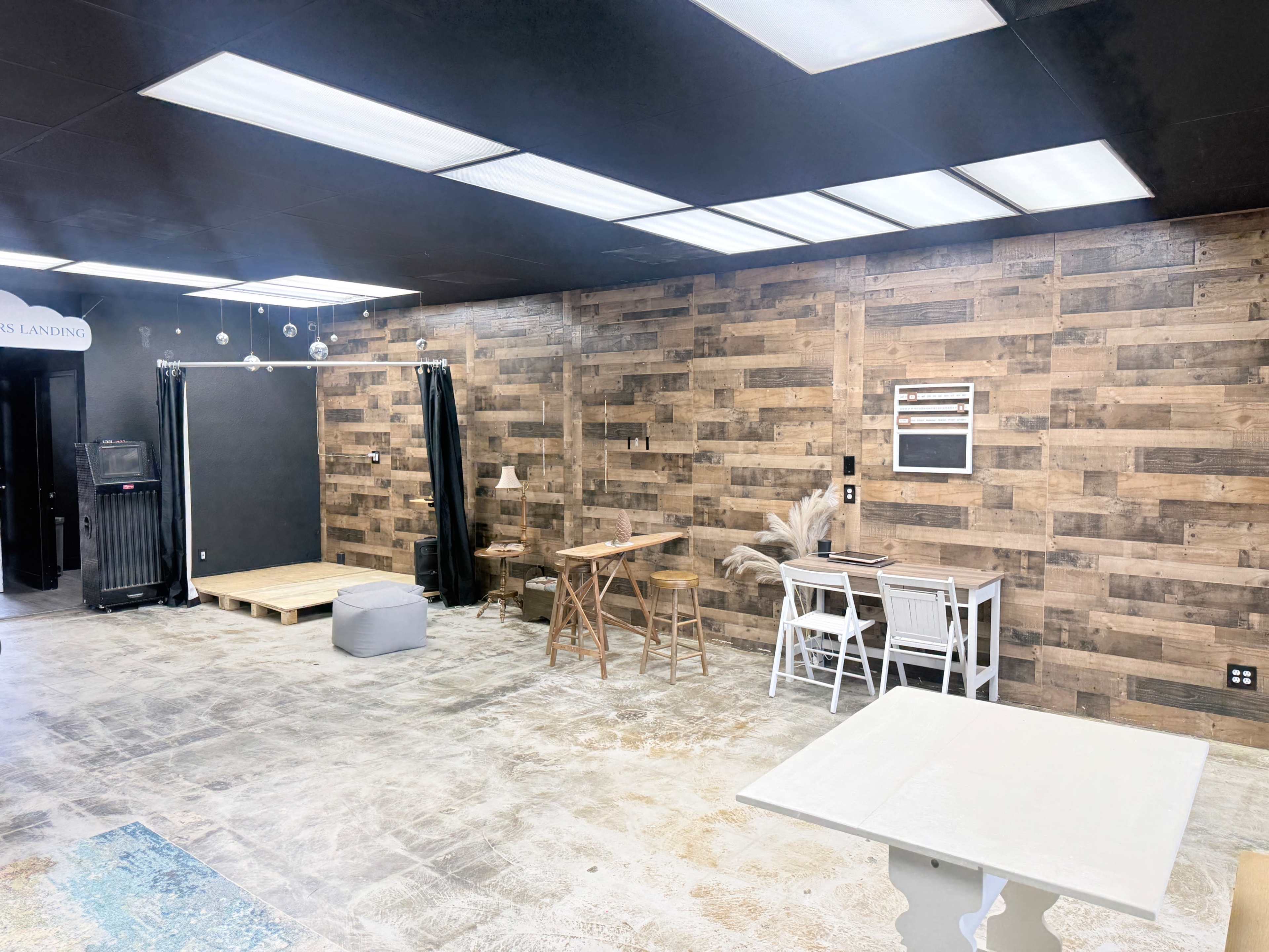 The image shows an empty, modern indoor space with wooden paneling on the walls, various tables, and some seating arrangements.