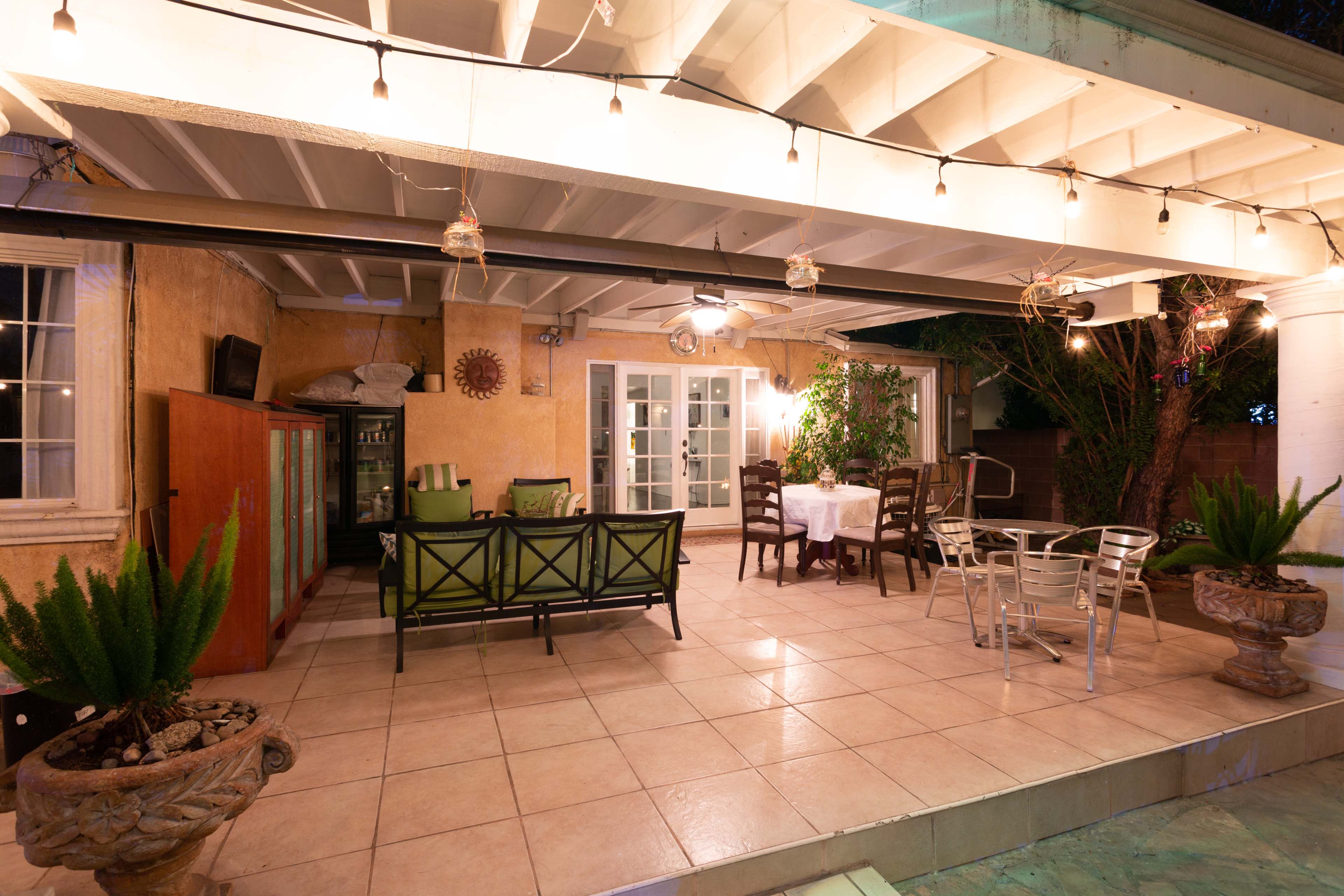 The scene shows a covered patio area with furniture, potted plants, and string lights.