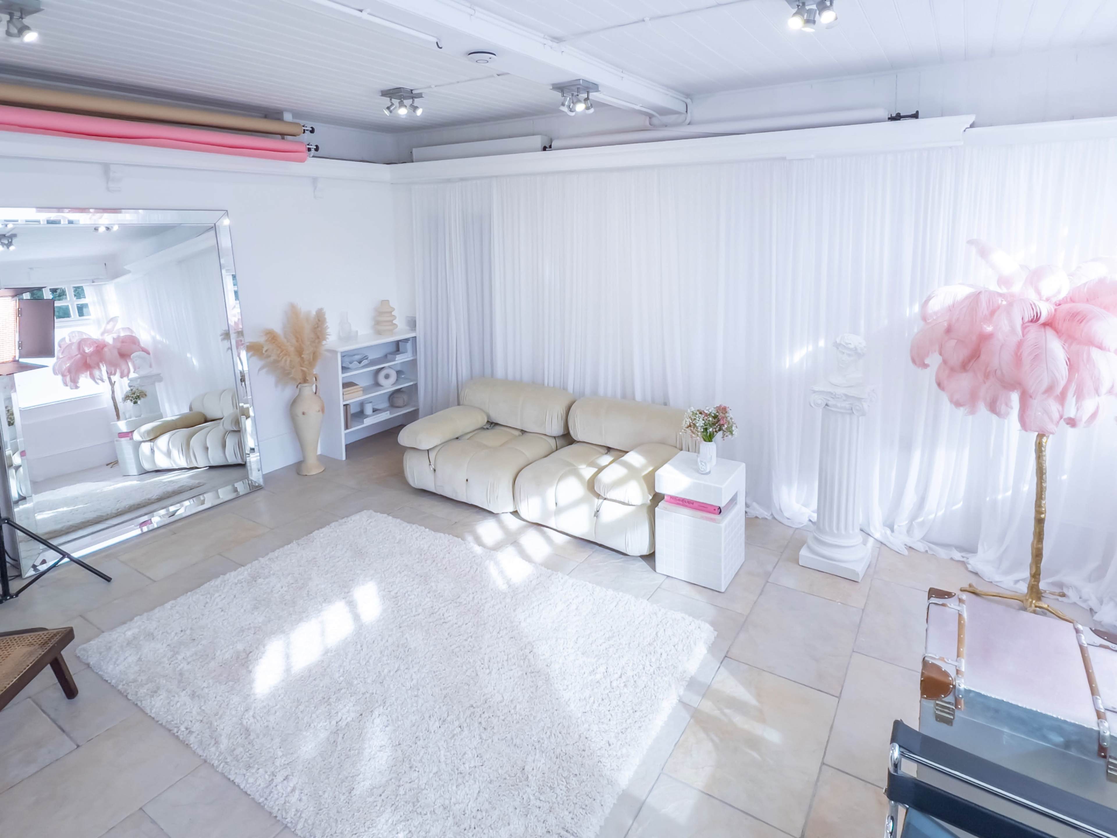A bright, minimalist studio space featuring a light-colored sofa, a fluffy area rug, a large mirror, and decorative elements such as a pink feather lamp and a white statue.