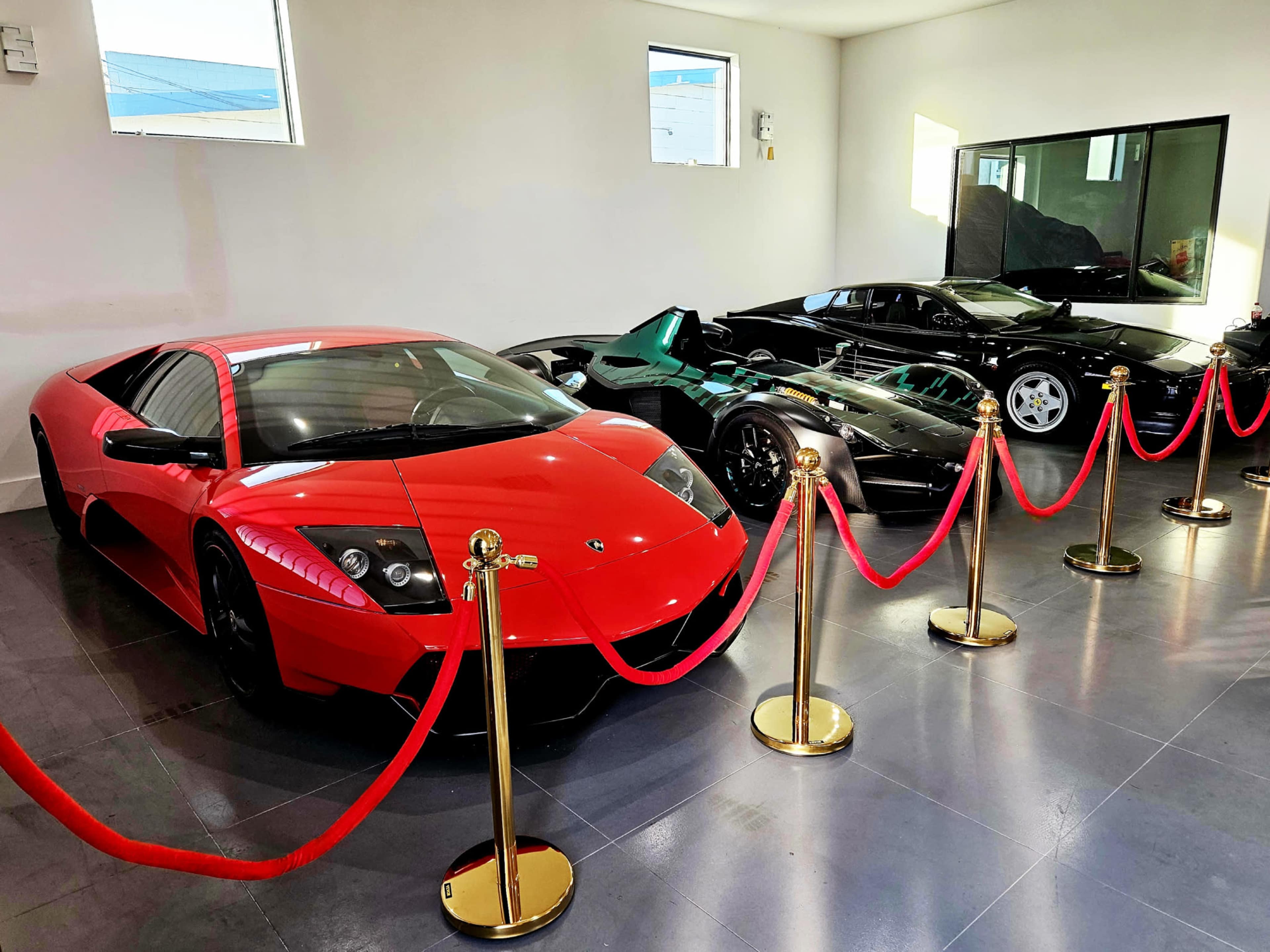 The image shows a modern showroom featuring a red Lamborghini Murciélago, a black McLaren P1, and a black Ferrari, all surrounded by red ropes and gold stanchions.
