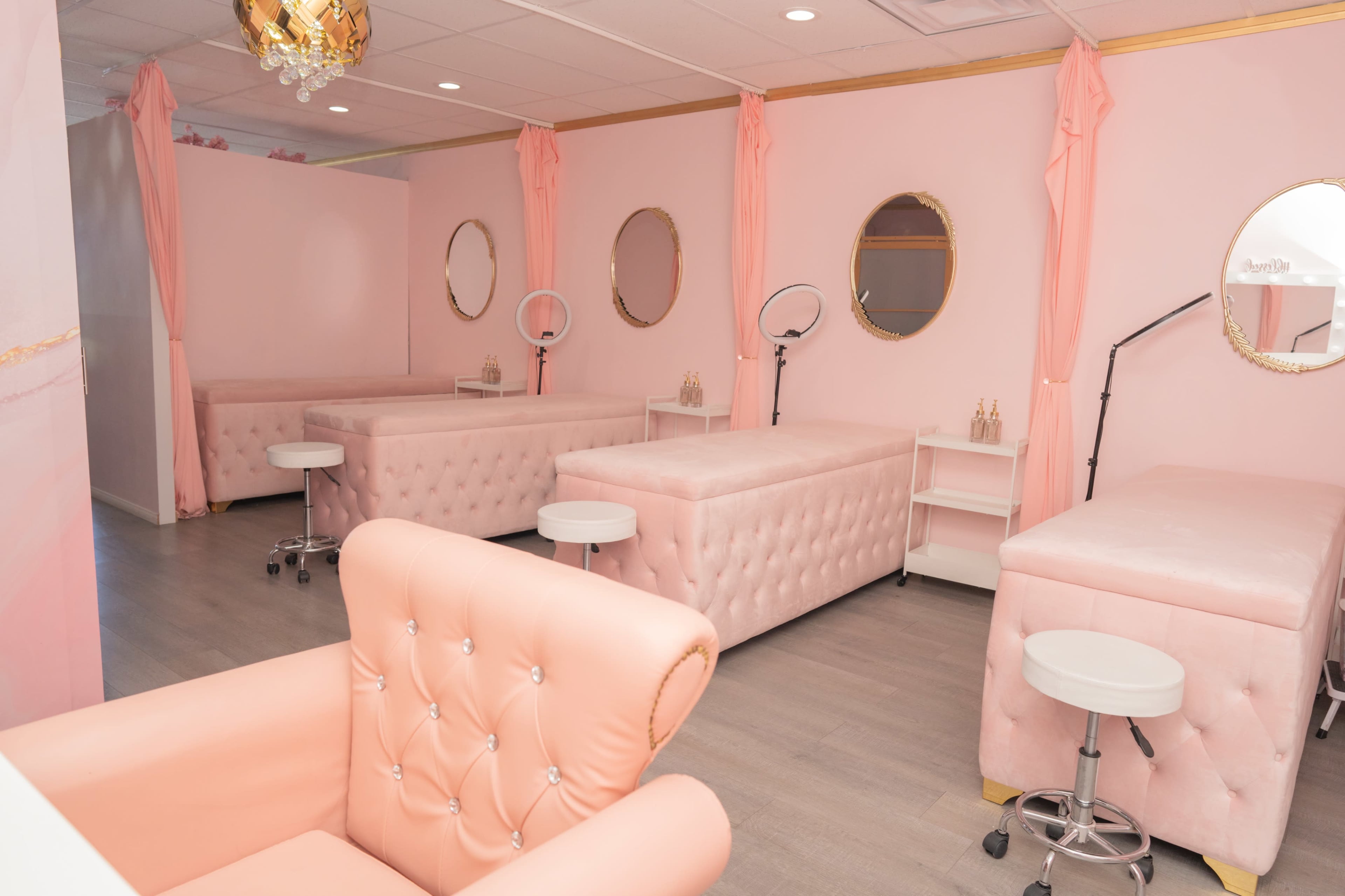 A beauty salon interior with pink upholstered treatment beds, mirrors, and styling equipment arranged in a spacious, well-lit room.