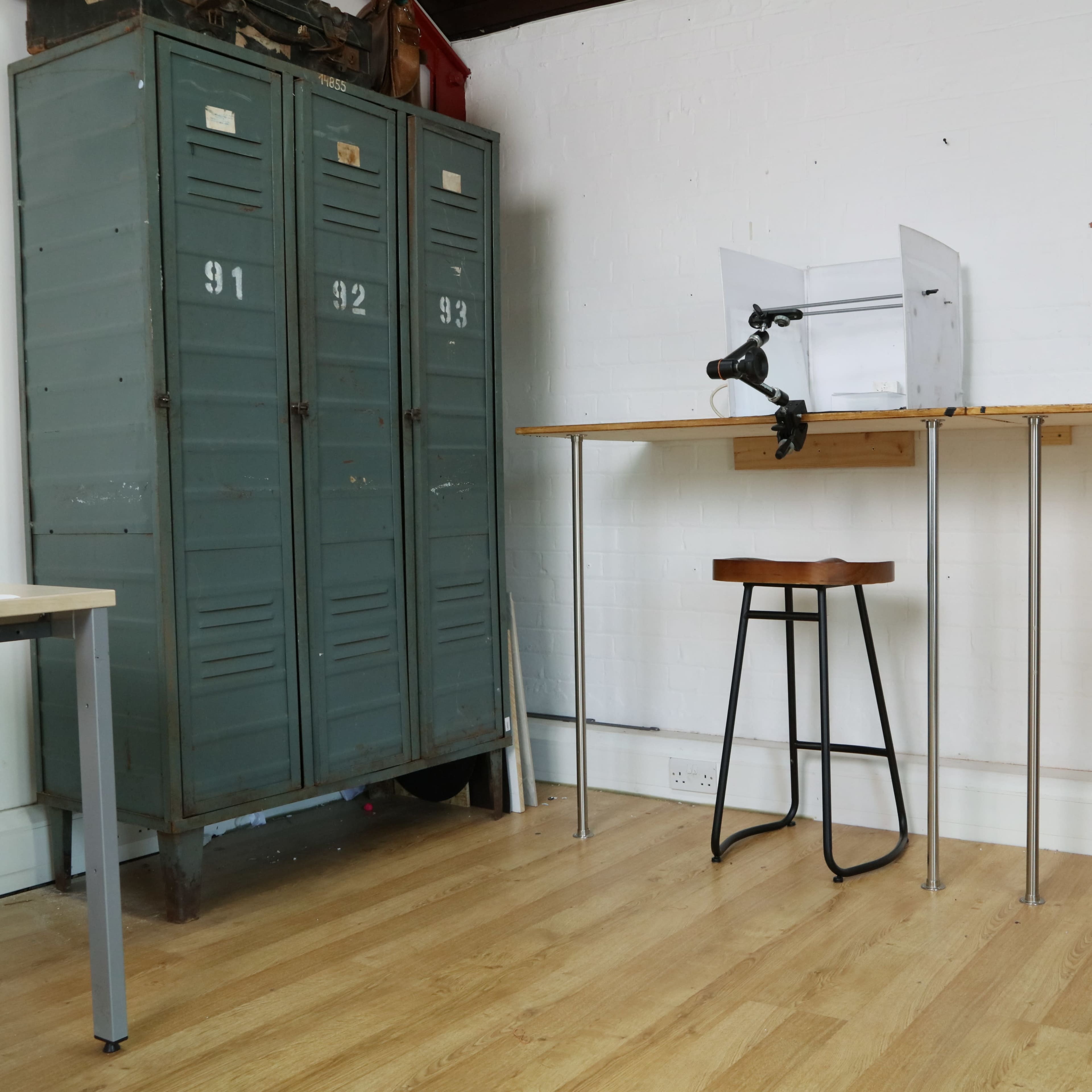 A minimalistic room featuring a pair of green lockers, a wooden table with a clamp and a stool, and light-colored wooden flooring.