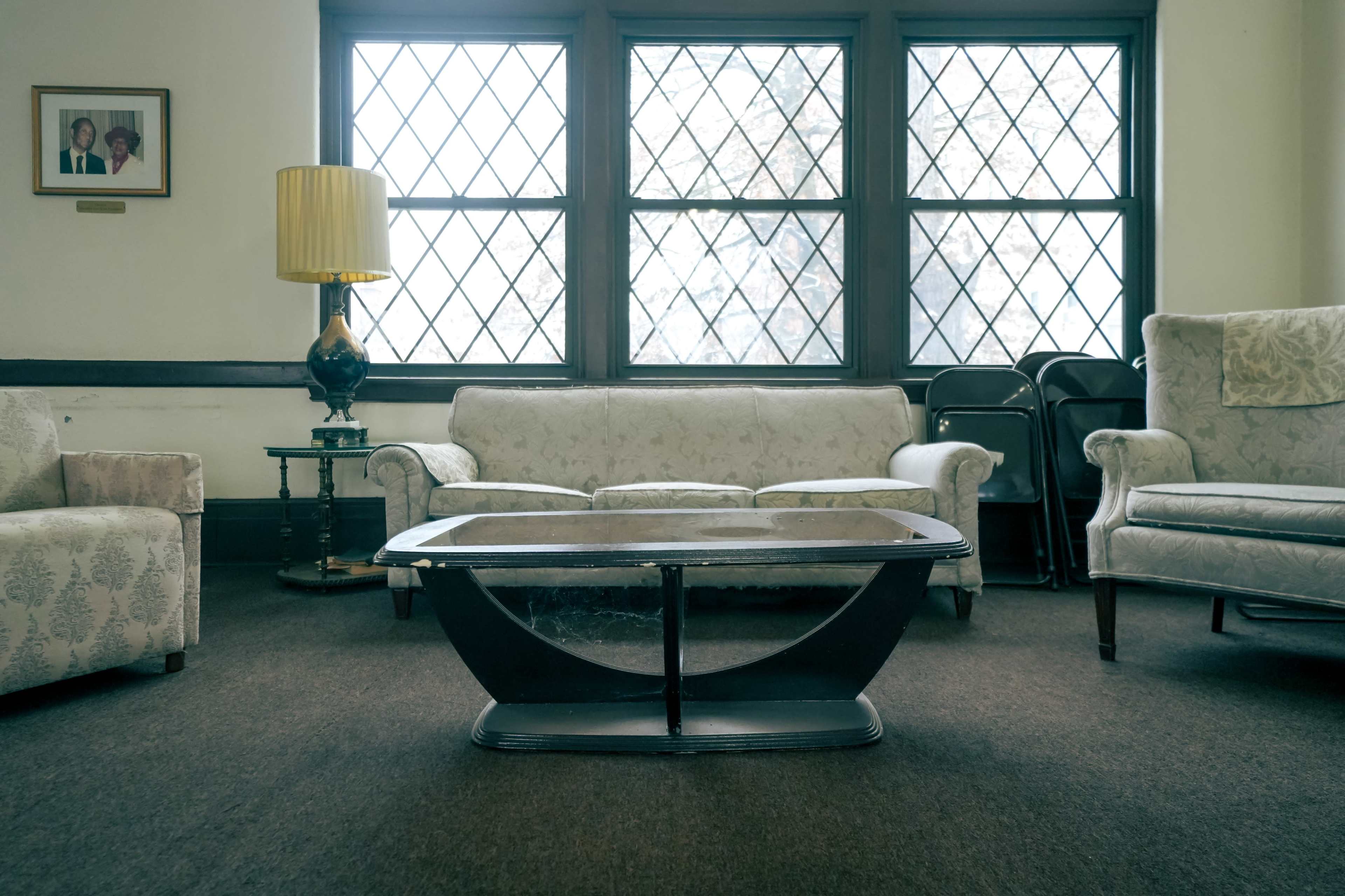 A room with two patterned sofas, a coffee table in the center, a lamp on a side table, and several folding chairs stacked against the wall by the windows.