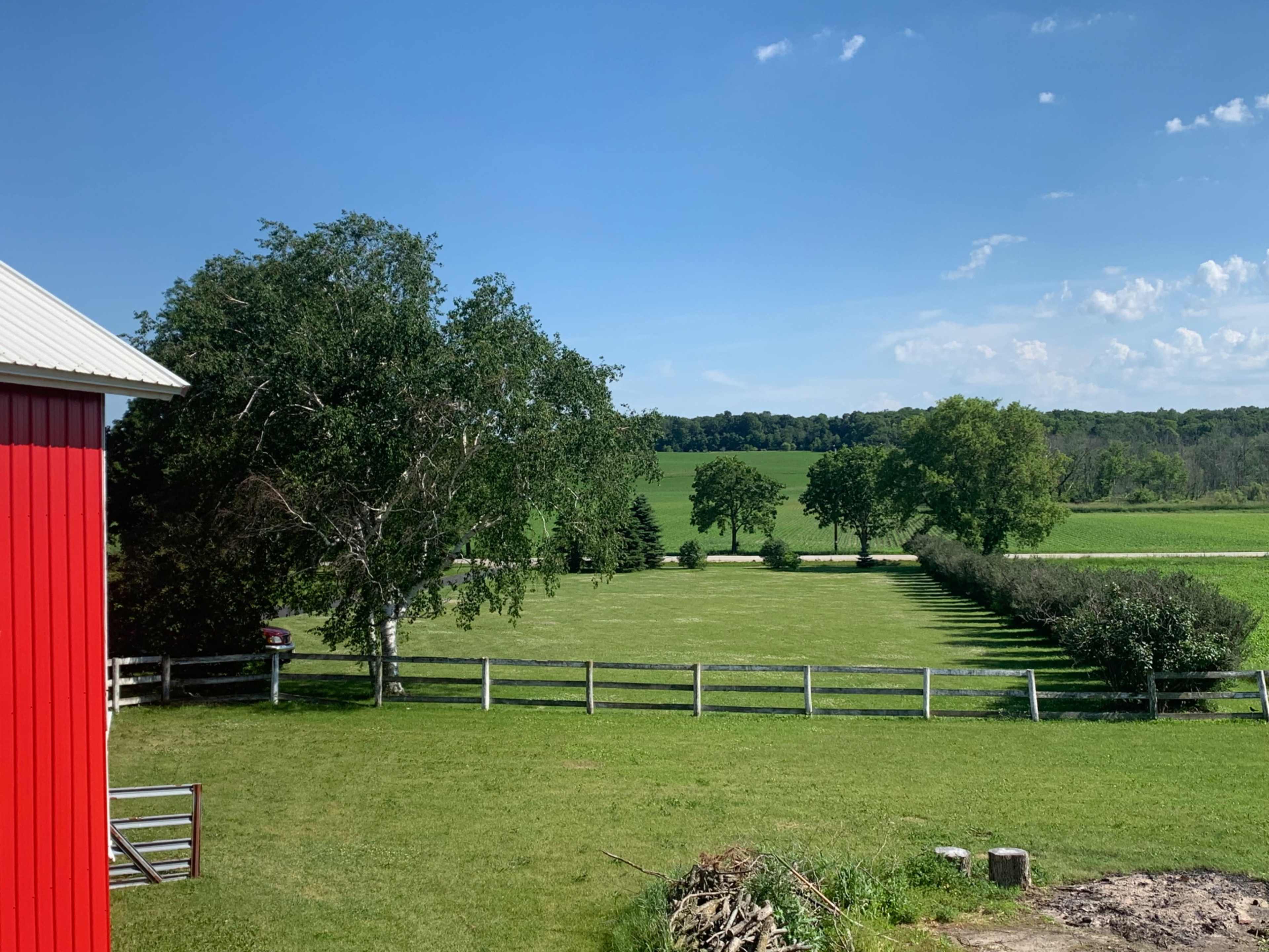 A red barn stands beside a green field lined with trees and a white fence under a clear blue sky.