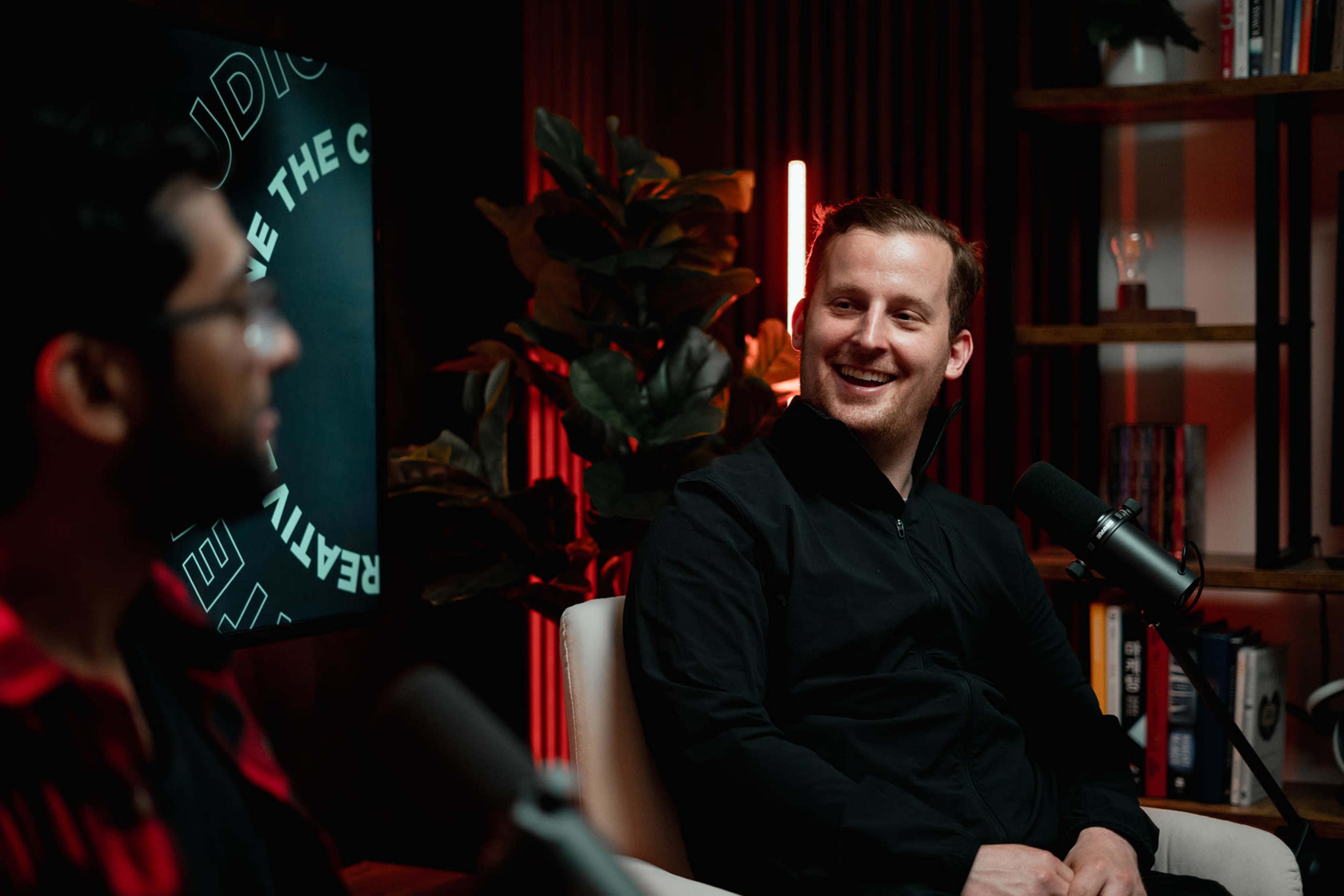 Two men are seated in a modern studio with a backdrop featuring plants and illuminated signage, engaged in conversation.