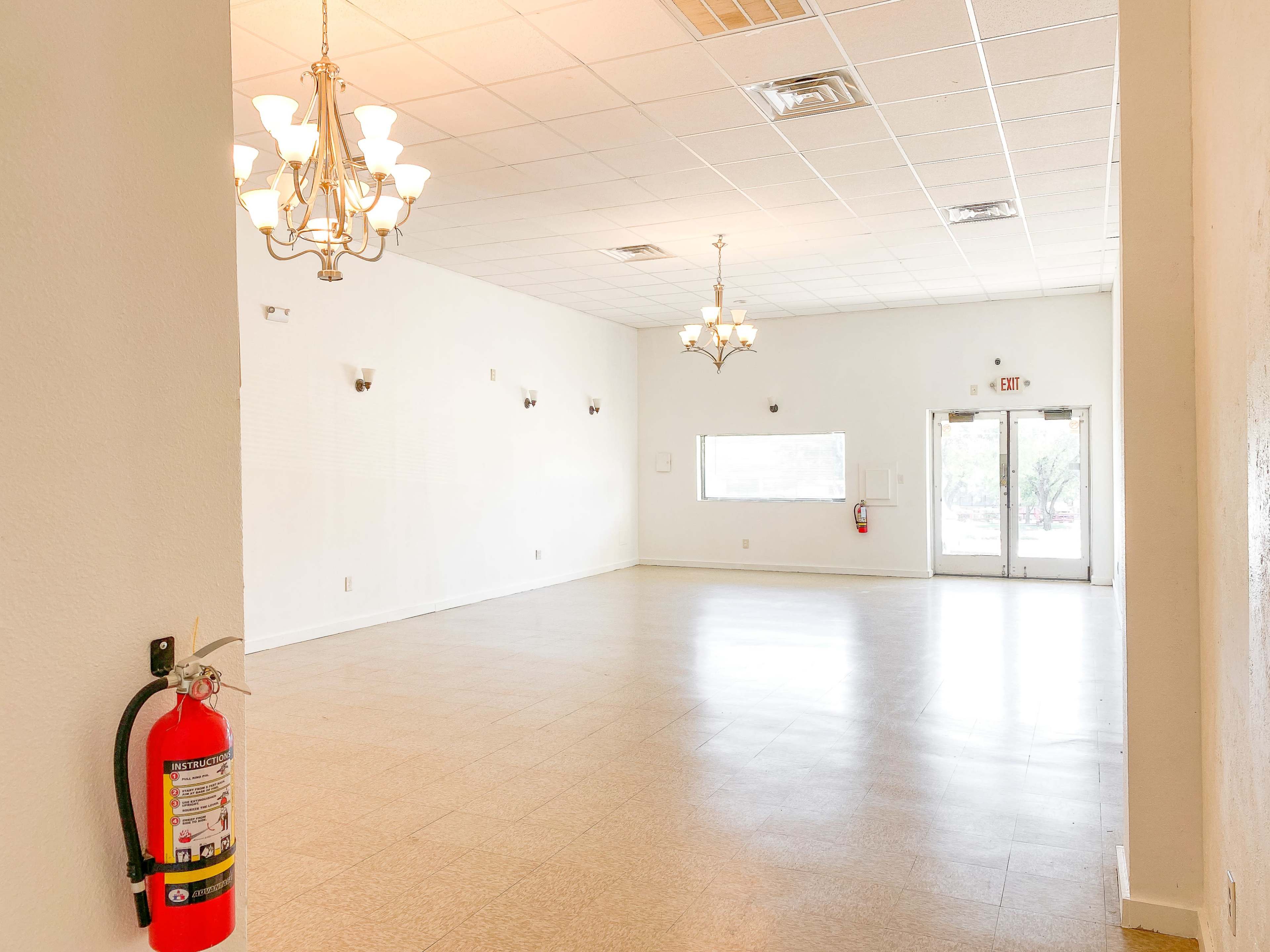 The image shows a spacious, empty room featuring two chandeliers and large windows with double doors leading outside.
