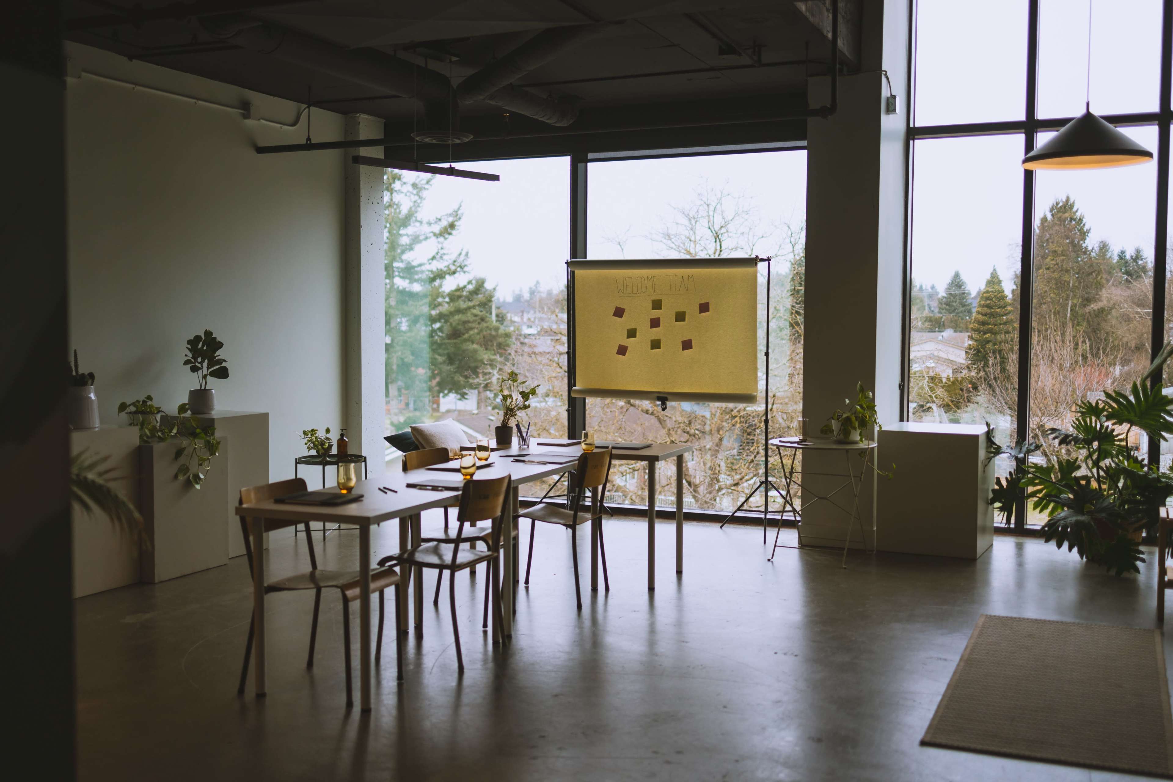 A spacious meeting room features a wooden table surrounded by chairs, with a presentation board and large windows overlooking a landscape of trees.