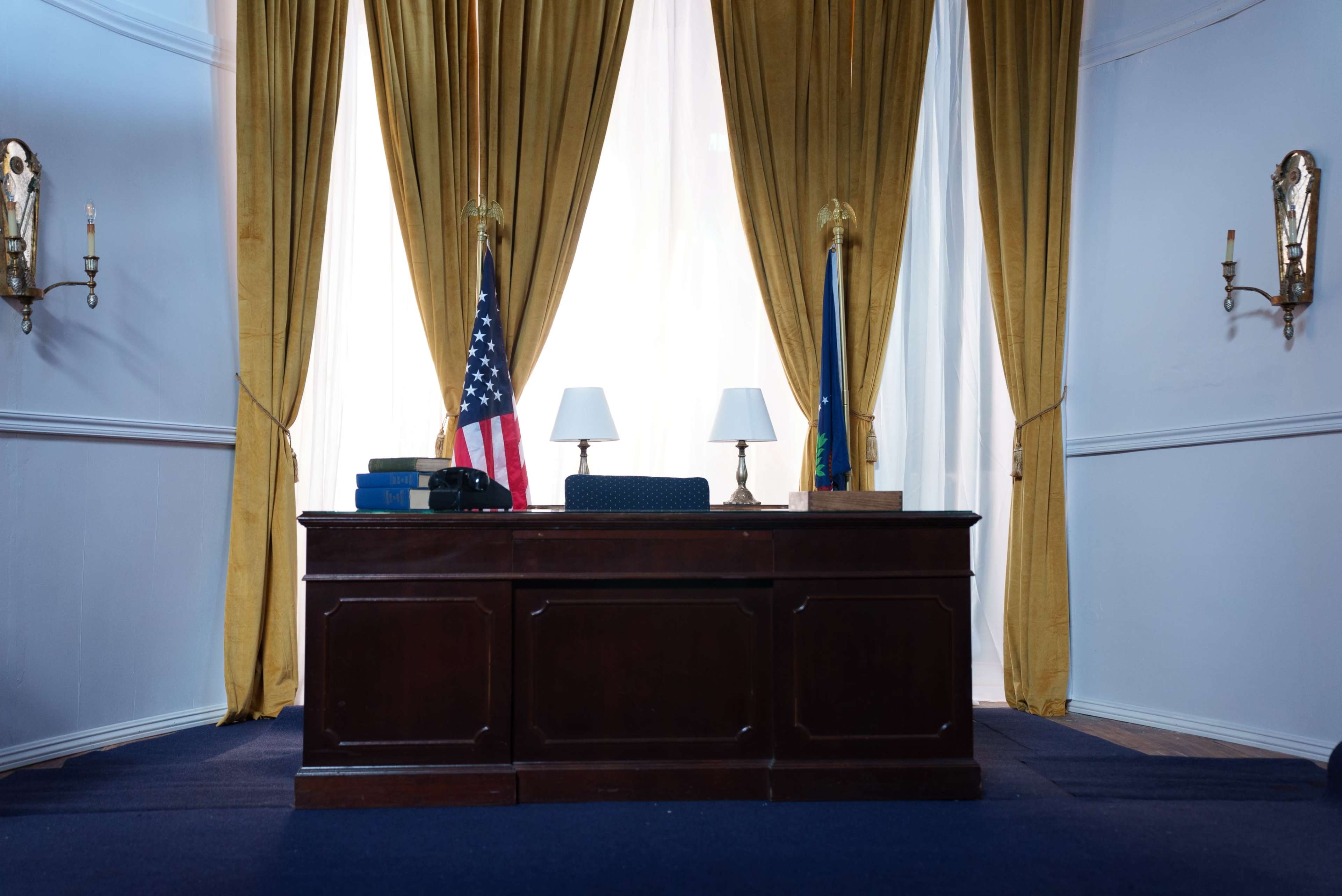 A wooden desk is positioned in front of a pair of golden draped curtains, with an American flag and a state flag on either side, and a lamp on the desk.