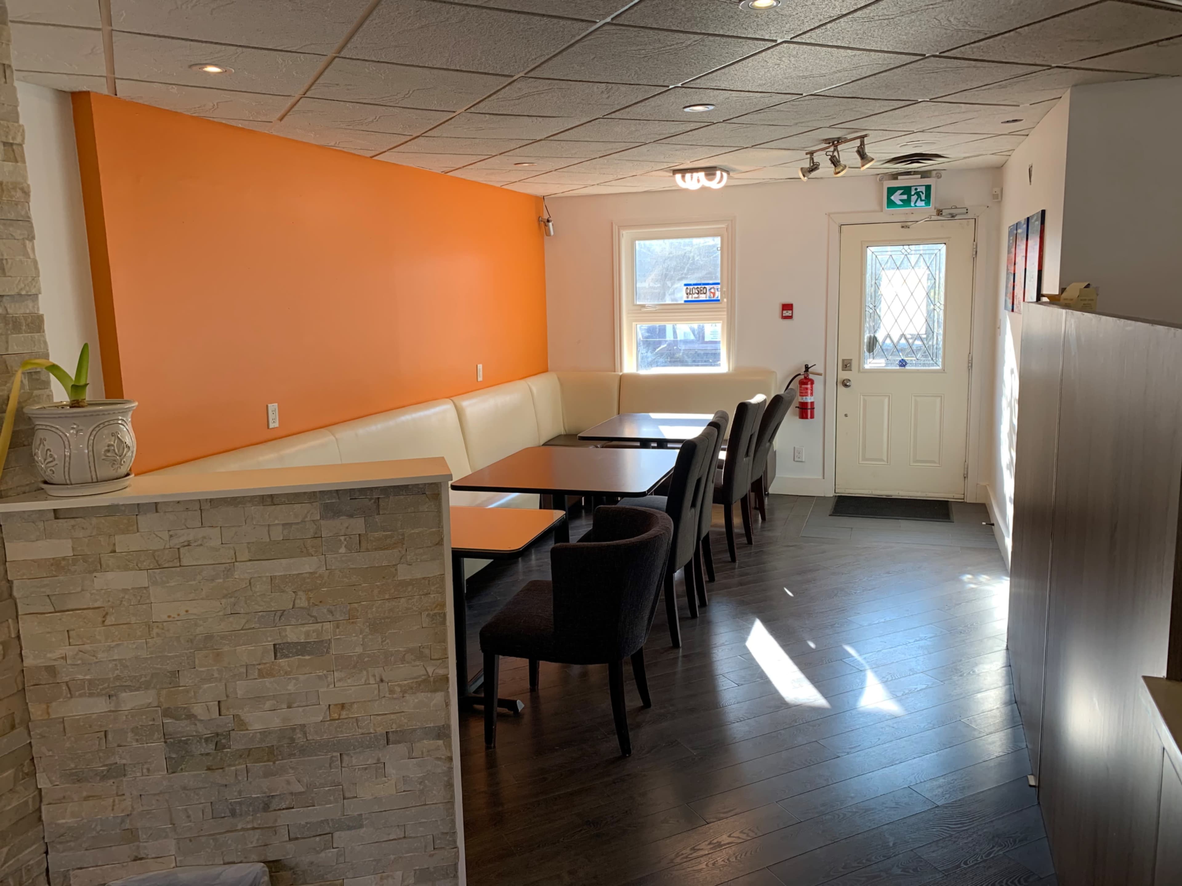 A small restaurant interior with a beige and orange color scheme, featuring a row of tables and chairs along one side and a large window near the entrance.