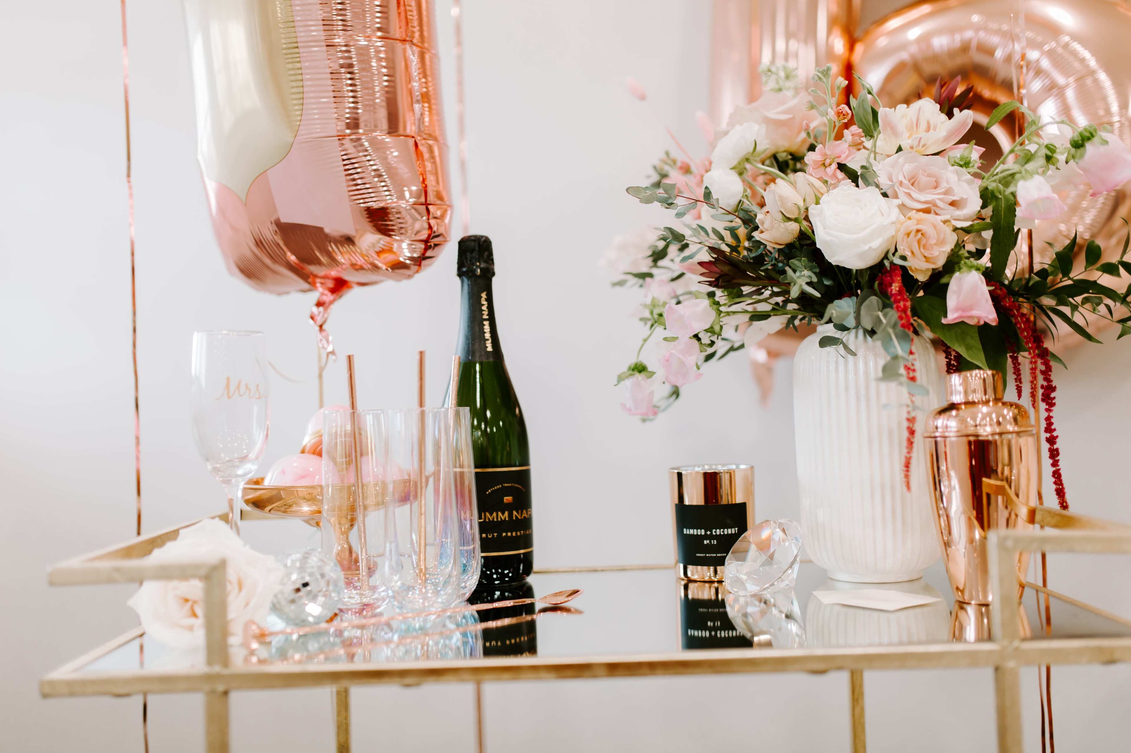 A bar cart is decorated with a bottle of champagne, elegant glasses, a floral arrangement, and rose gold balloons.