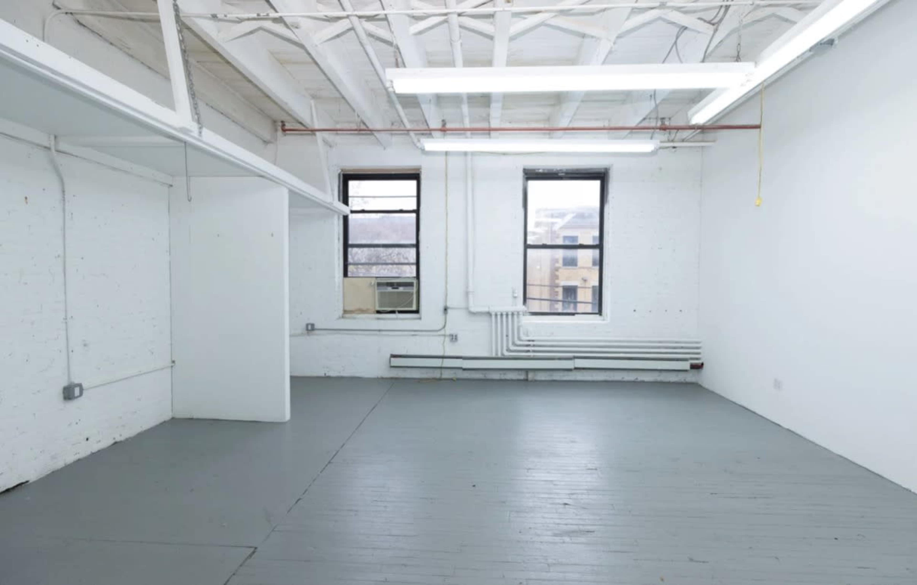 The image shows an empty room with white walls, a wooden floor painted gray, and two windows on one side.