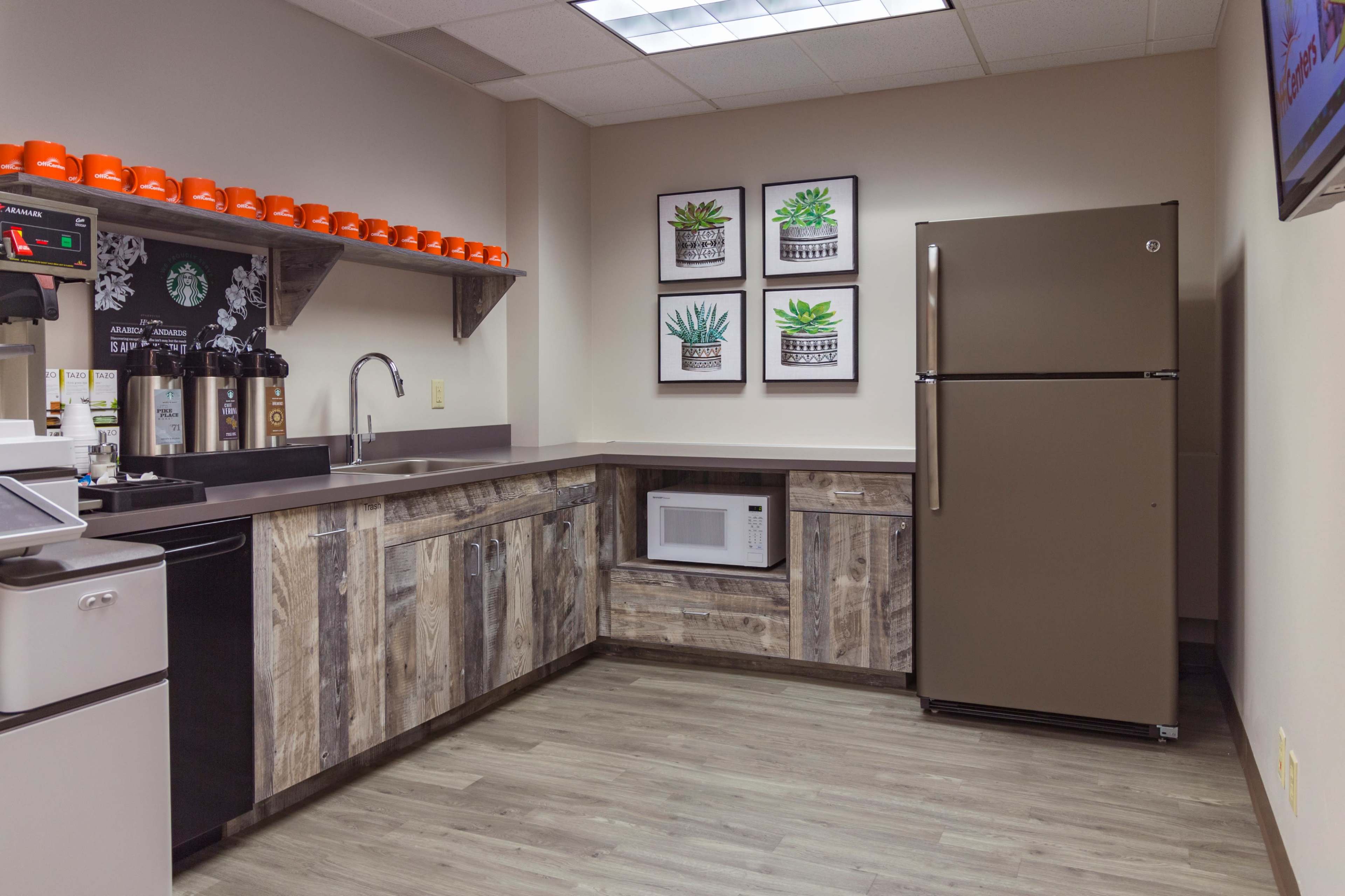 A modern kitchen area featuring a refrigerator, microwave, and coffee station, with wooden cabinetry and framed plant artwork on the wall.