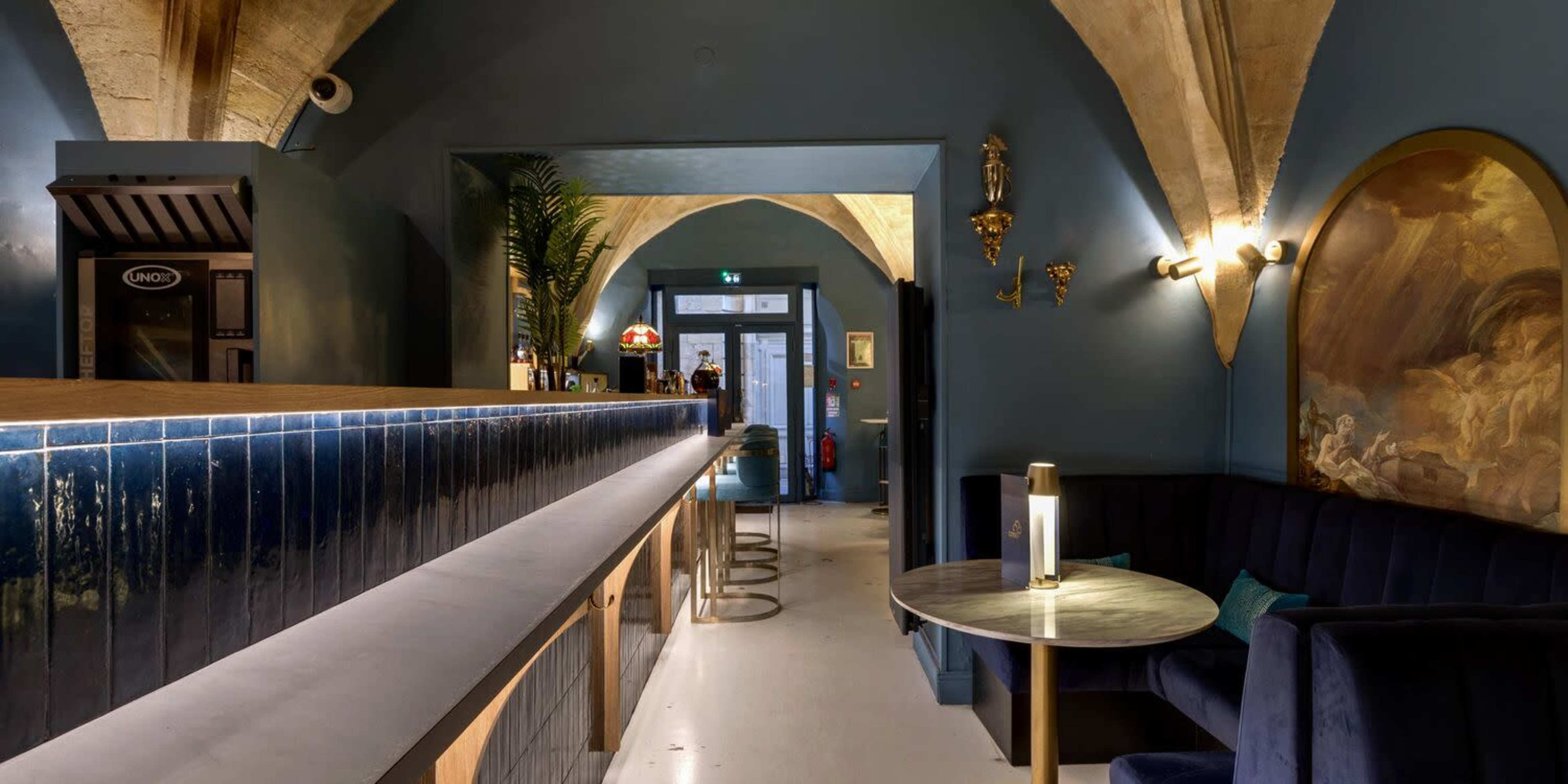 The image shows a modern bar interior with blue walls, arched ceilings, and a long countertop, featuring a lounge area with plush seating.