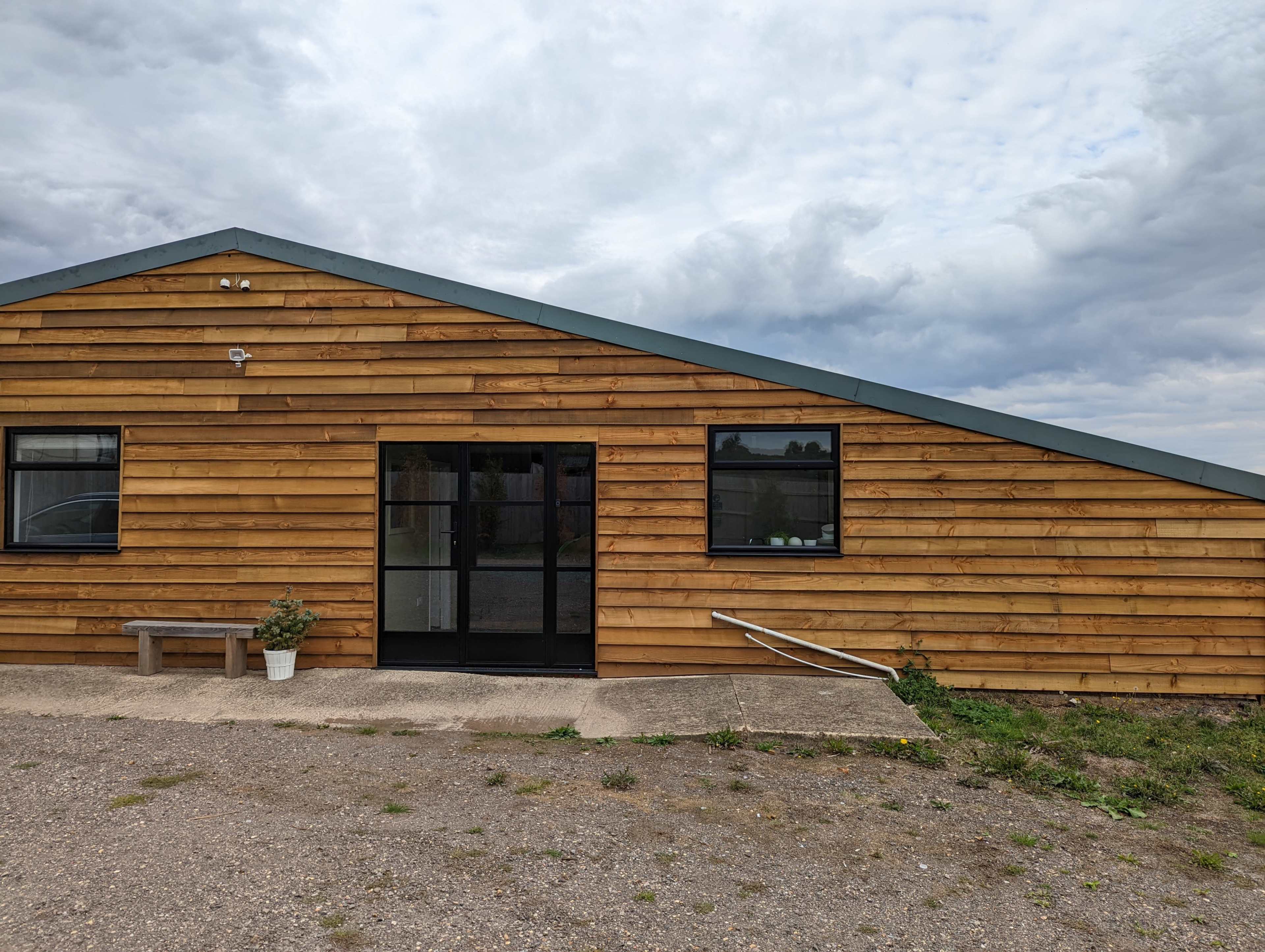 A wooden building with a slanted roof, a glass door, and windows, situated on a gravel lot under a cloudy sky.