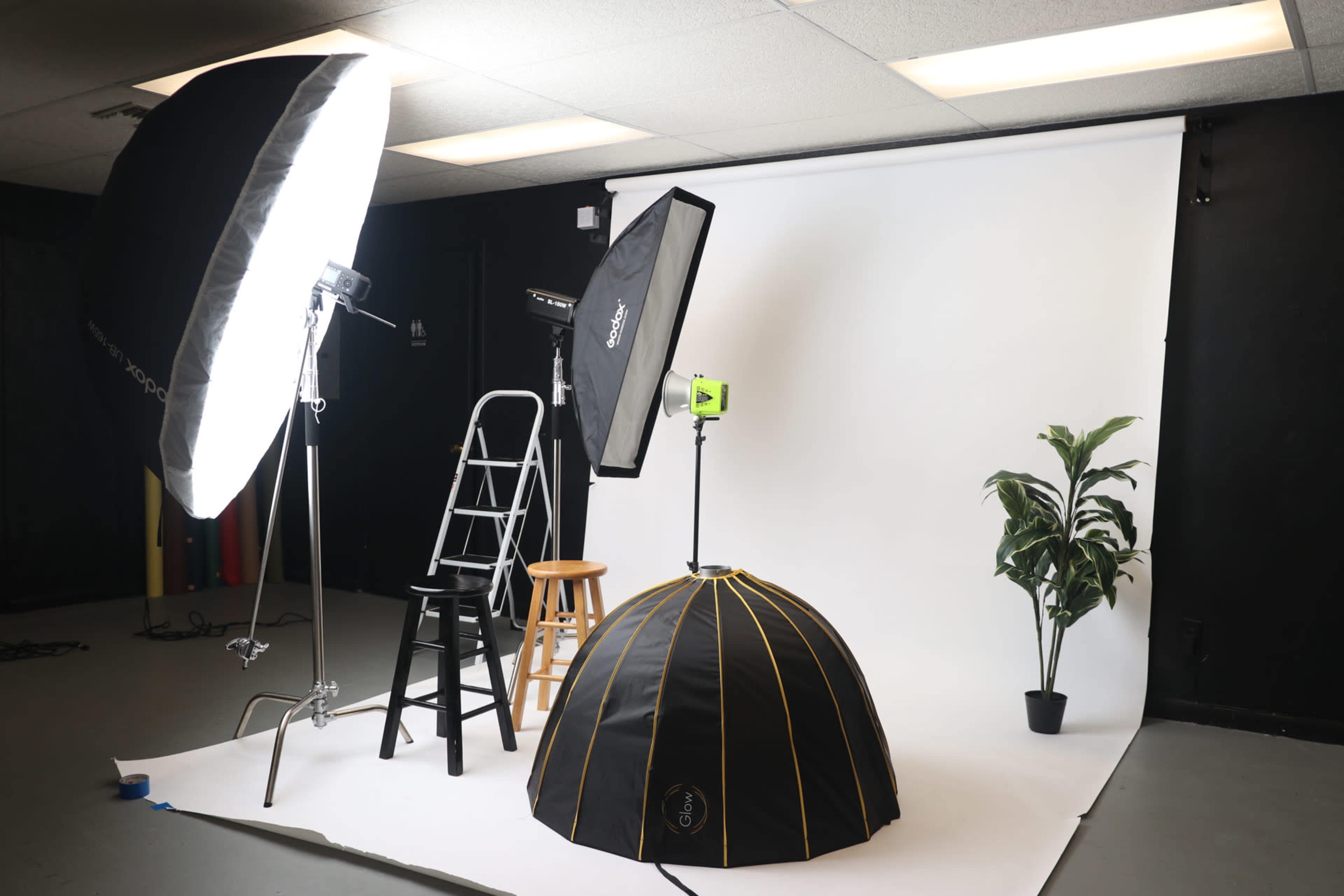 A photography studio setup featuring a white backdrop, various lighting equipment, a ladder, a black stool, a plant, and a light modifier.