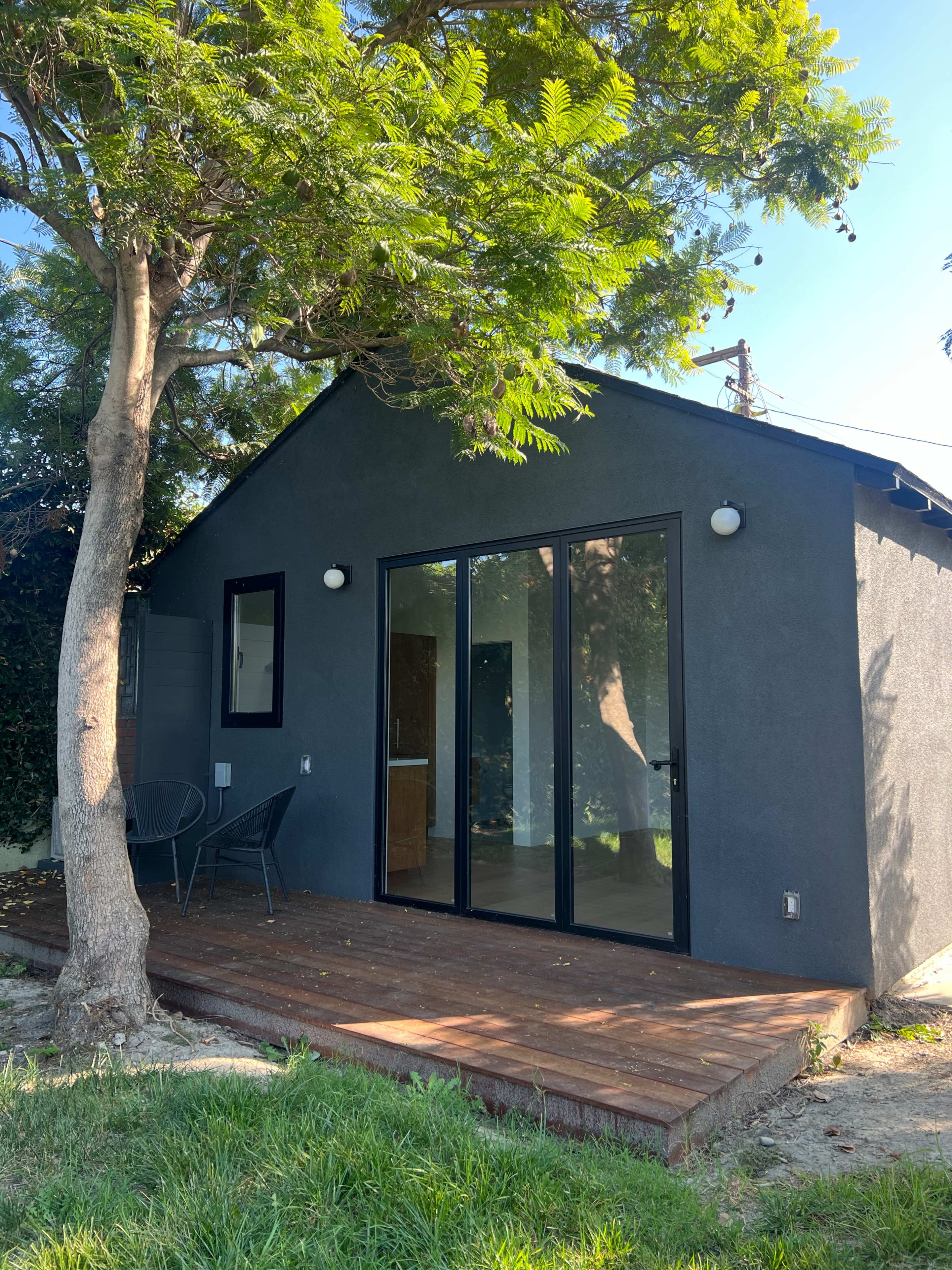 A small black house with large glass doors and a wooden deck is surrounded by greenery.